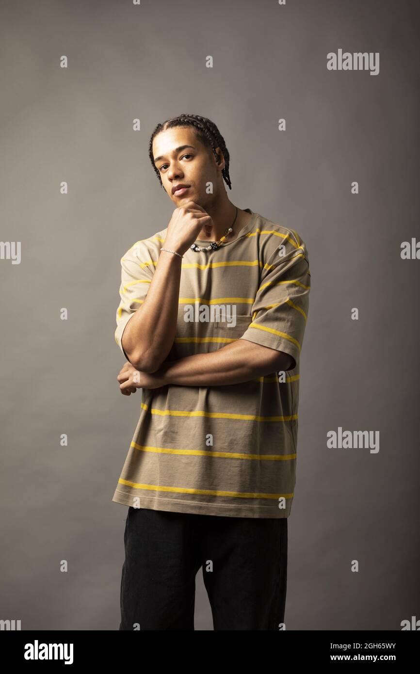 Jeune Afro-américain réfléchi modèle masculin avec des cheveux tressés vêtu d'une chemise et d'un collier à larges rayures regardant l'appareil photo contre le dos gris Banque D'Images