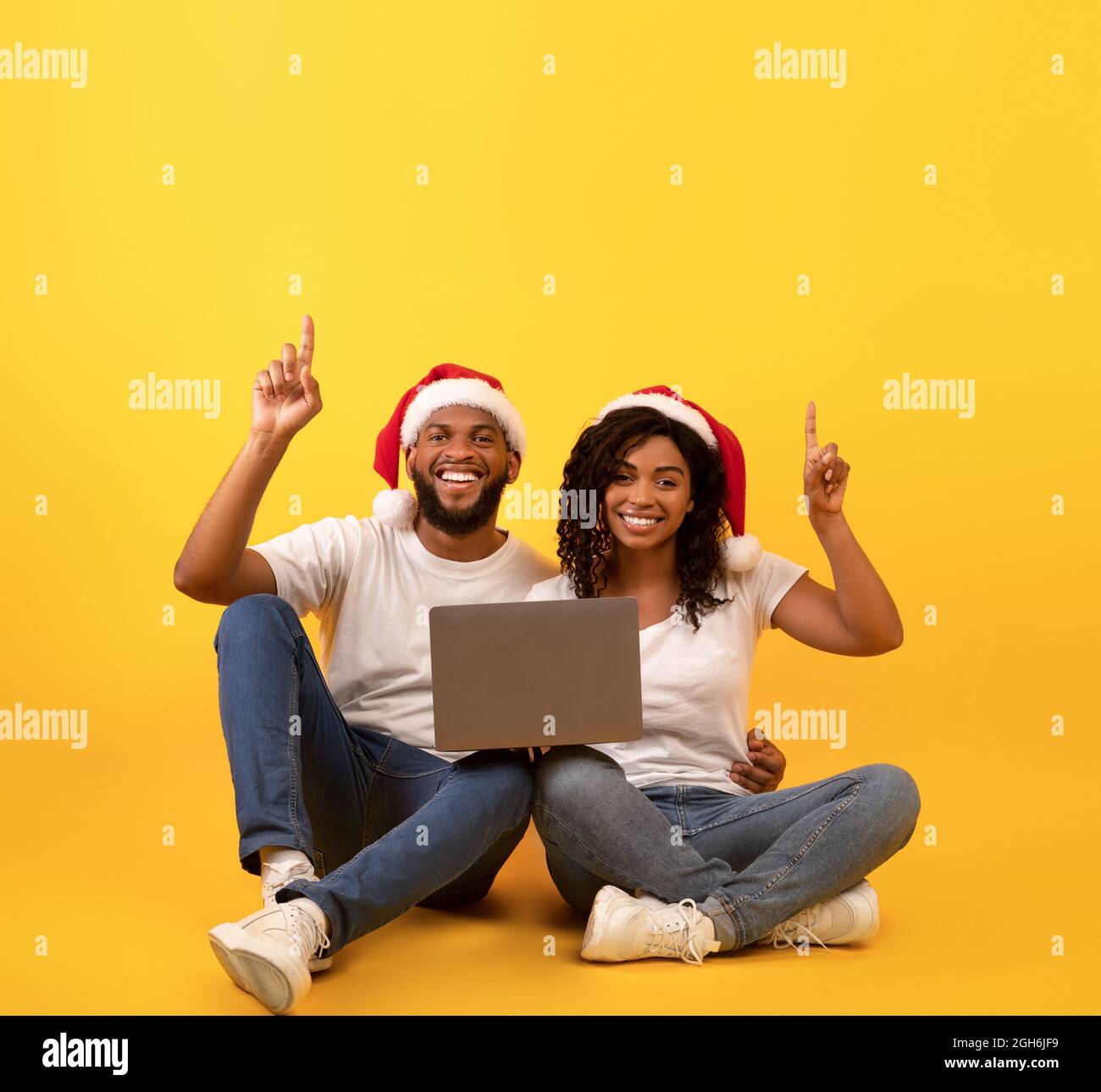 Offre de Noël. Heureux couple afro-américain assis avec un ordinateur portable et pointant vers l'espace libre, fond jaune Banque D'Images
