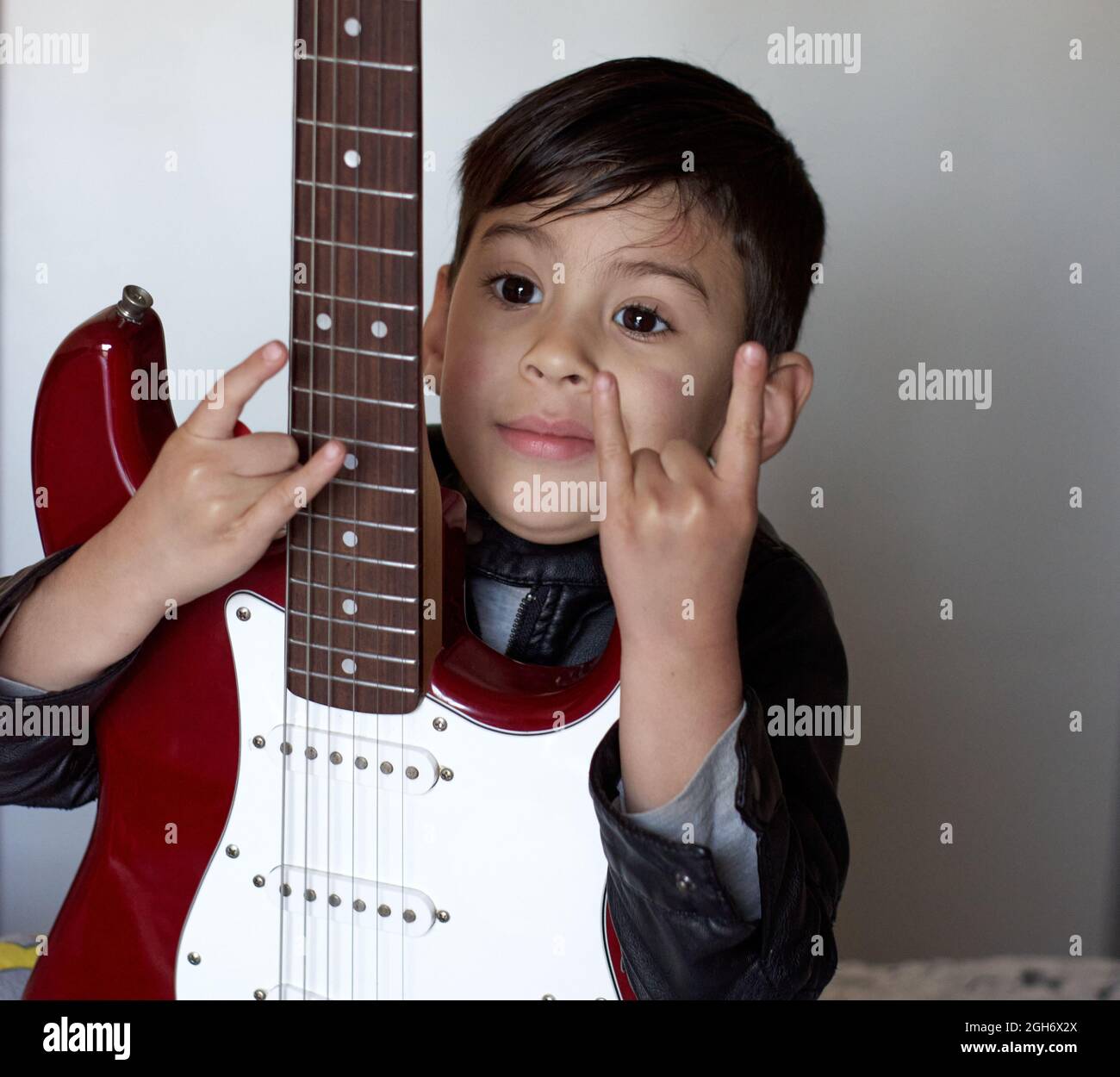 Un petit garçon hispanique montre des signes Rock and Roll et tient une guitare électrique rouge isolée sur fond blanc. Signe des avertisseurs sonores Banque D'Images