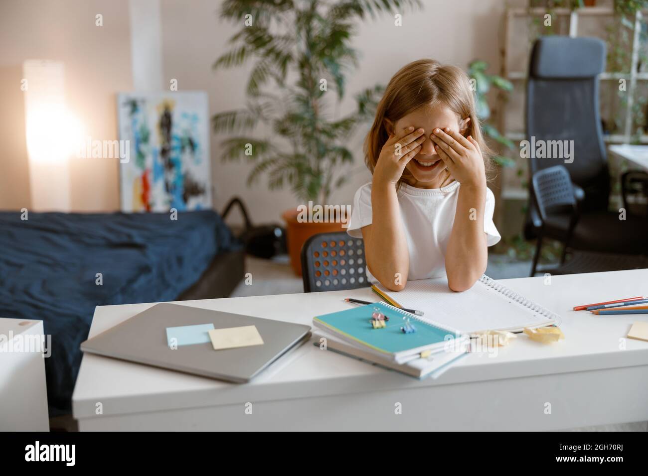 Un petit étudiant enjoué ferme les yeux près des paumes du bureau avec des fournitures et un ordinateur portable à la maison Banque D'Images