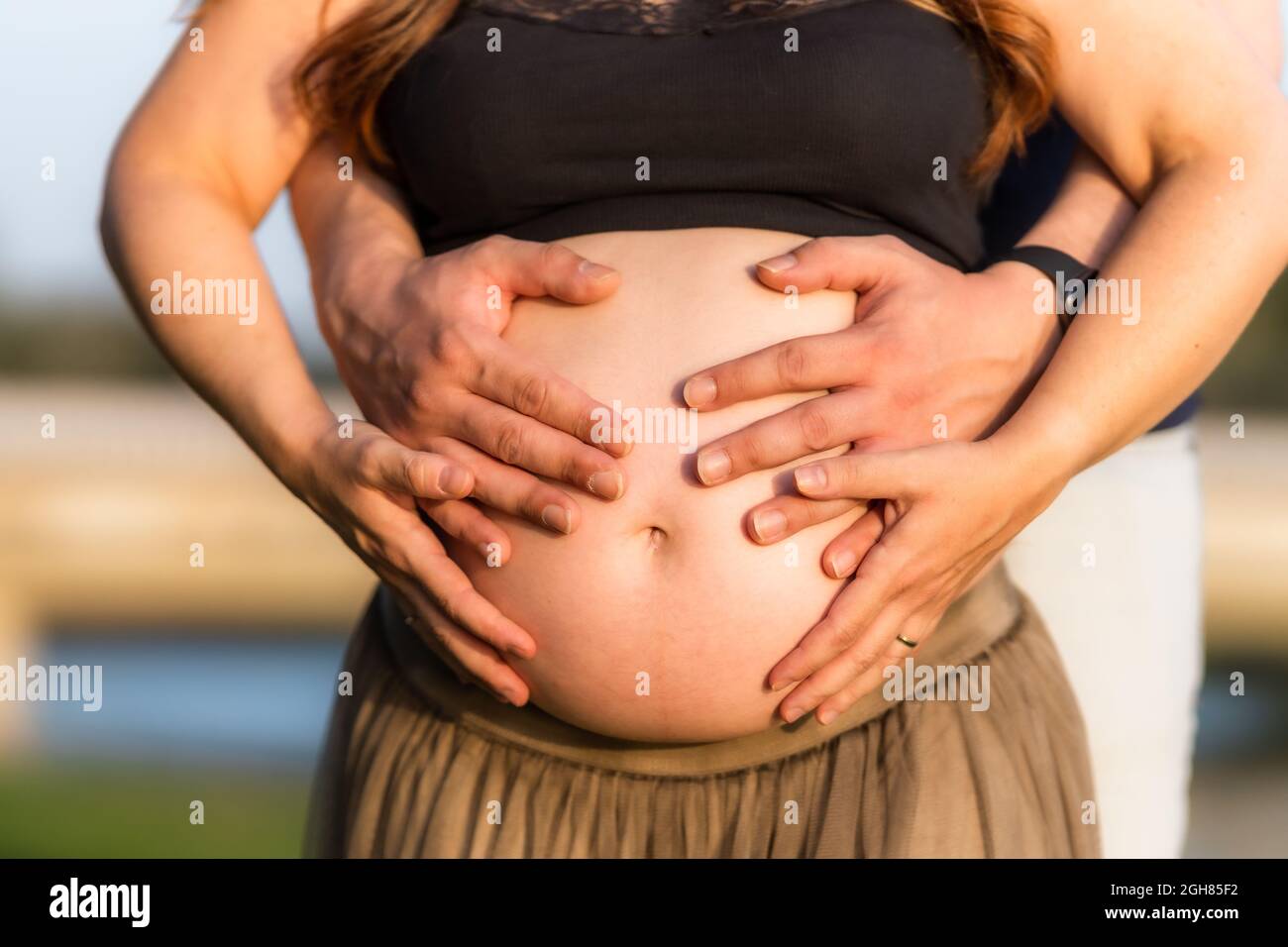 Vue rapprochée de quatre mains sur l'abdomen d'une femme enceinte adulte debout à l'extérieur Banque D'Images
