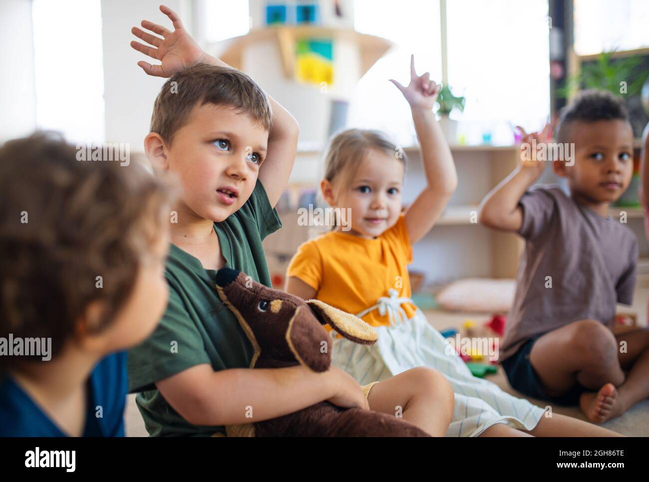 Groupe de petits enfants de la maternelle assis sur le sol à l'intérieur dans la salle de classe, levant les mains. Banque D'Images