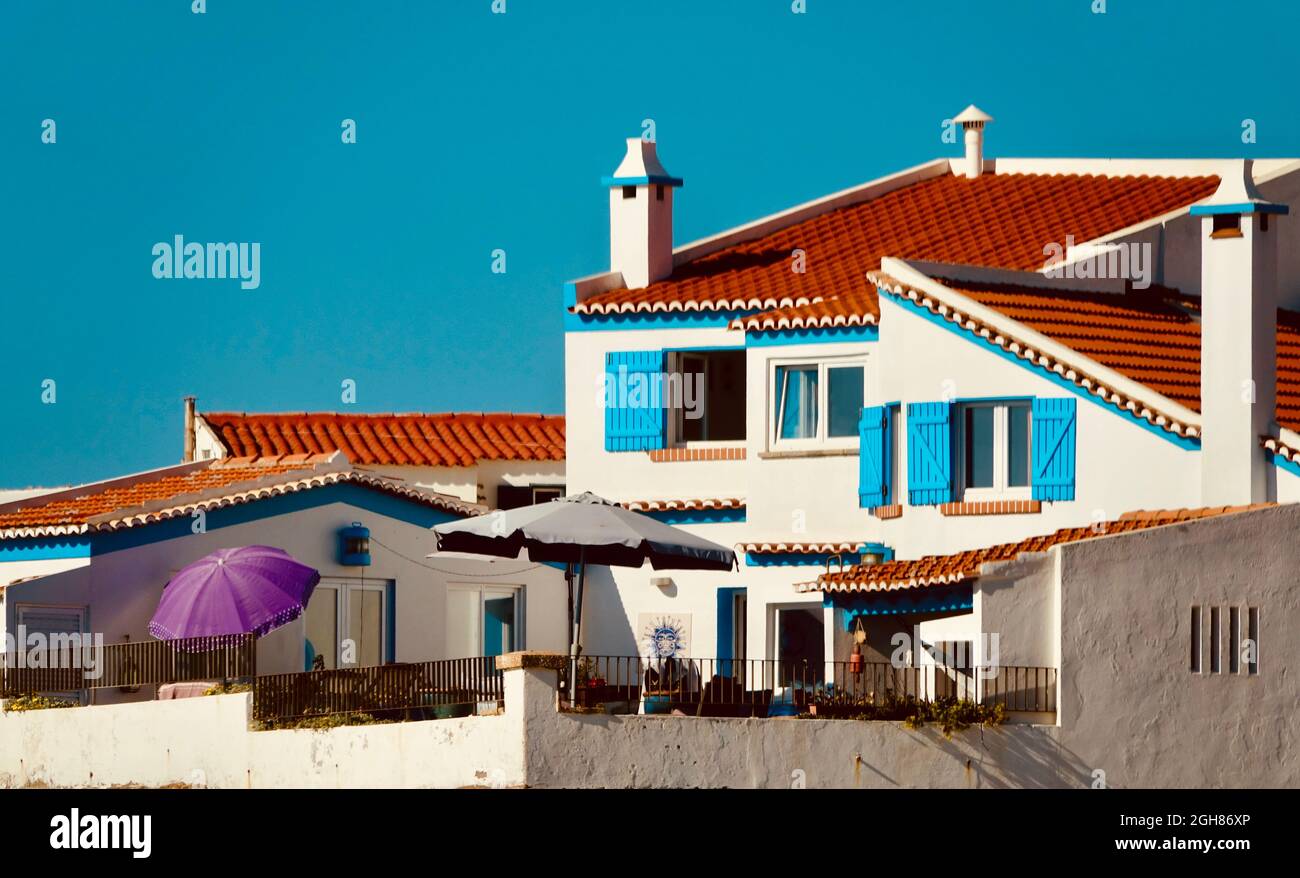 Vue sur une maison avec une façade et des cheminées blanches, des fenêtres avec volets bleus et un parasol violet, sur une falaise sur la côte portugaise Banque D'Images