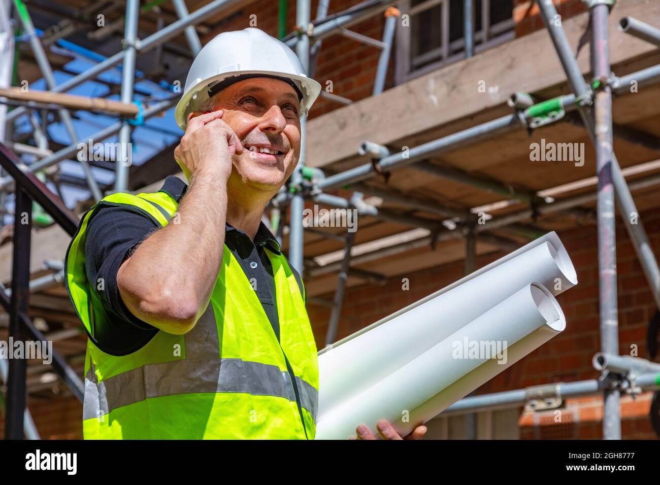 Homme constructeur contremaître, travailleur ou architecte sur le chantier de construction, heureux, souriant, tenant des plans de construction et parlant sur son téléphone cellulaire Banque D'Images