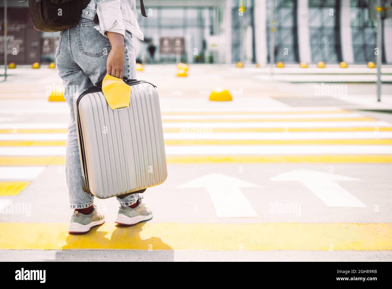 Femme voyageur traversant une route tenant une valise à bagages et un masque de protection jaune. Prêt pour un vol entrant dans l'aéroport, concept de voyage moderne Banque D'Images