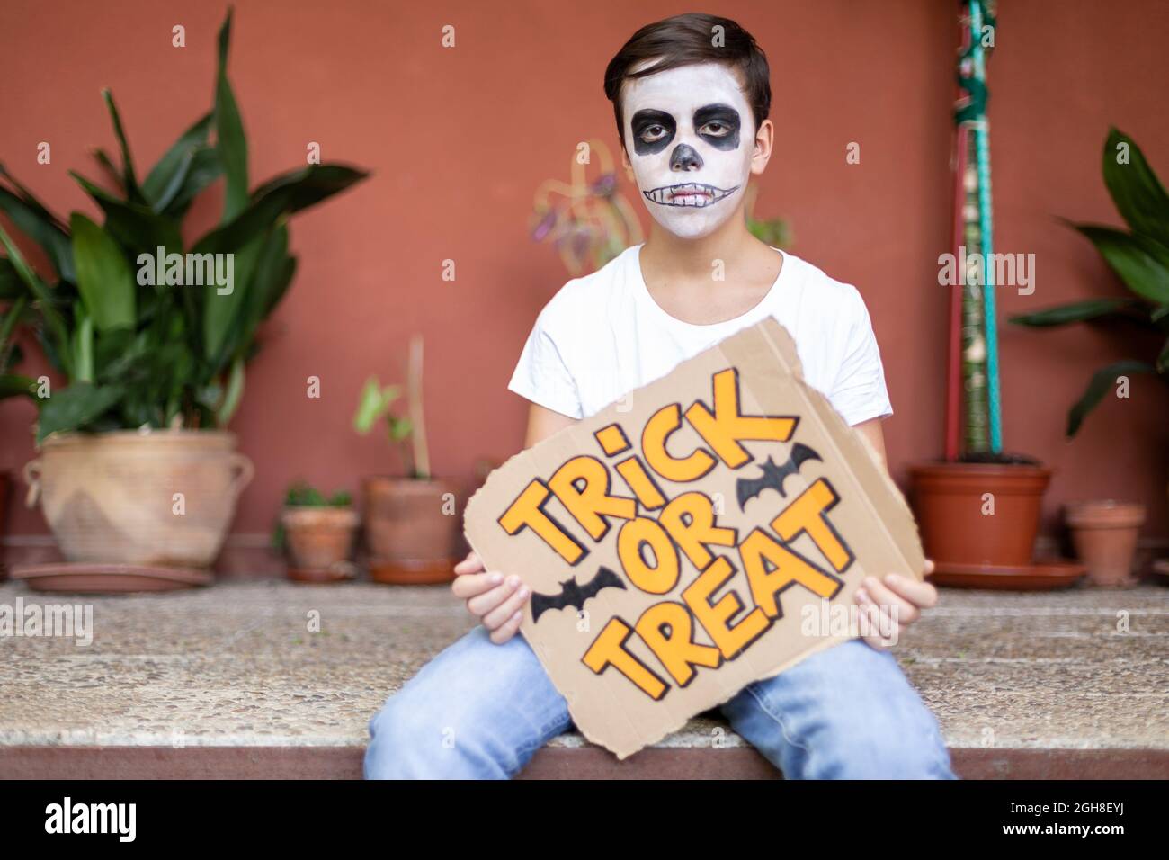 Adolescent à l'entrée de sa maison. Il a son visage composé et tient un signe avec le texte trick ou traiter. Halloween Celebration concept. Banque D'Images