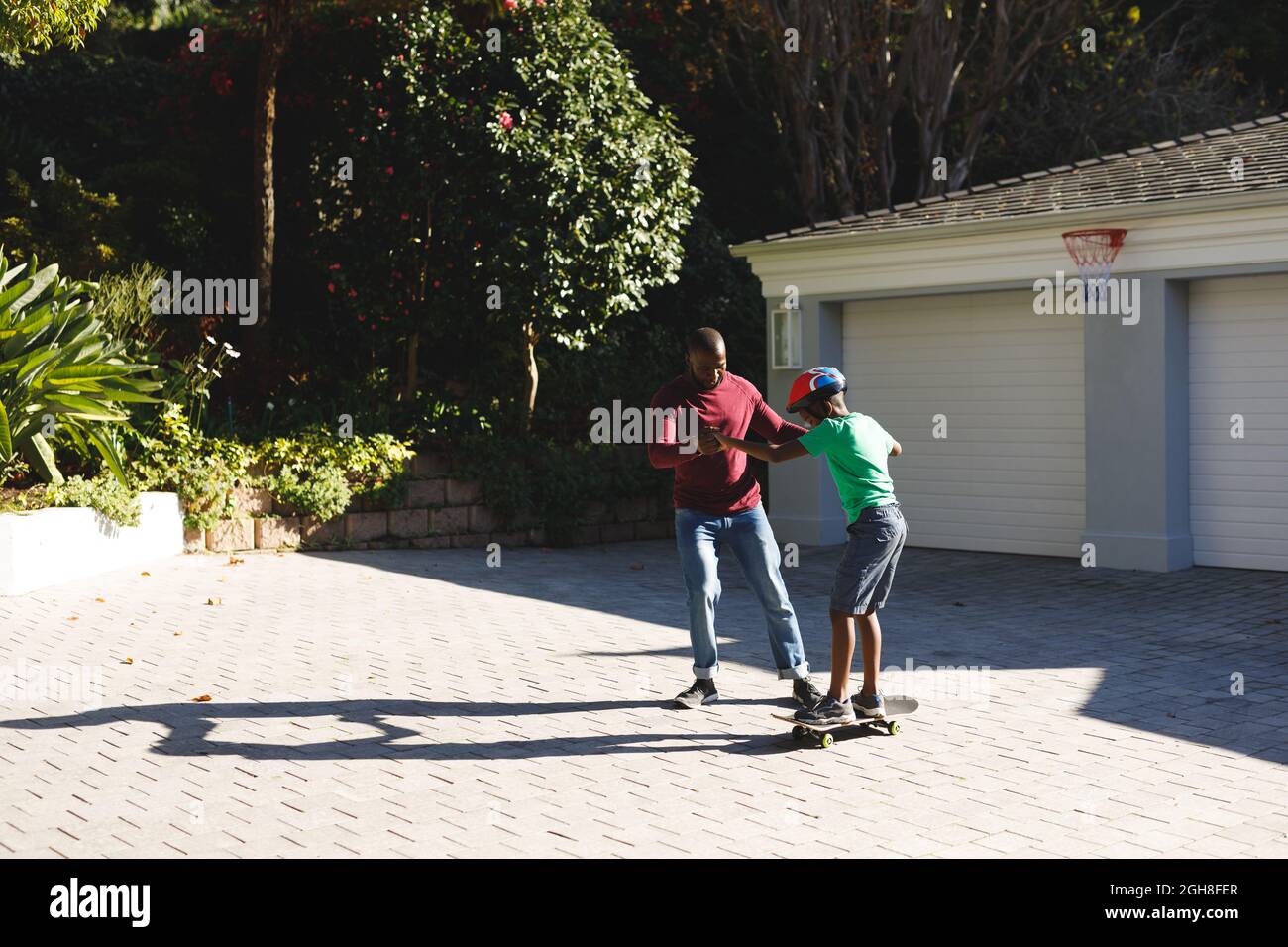 Père afro-américain souriant et aidant son fils à s'équilibrer sur le skateboard dans le jardin Banque D'Images