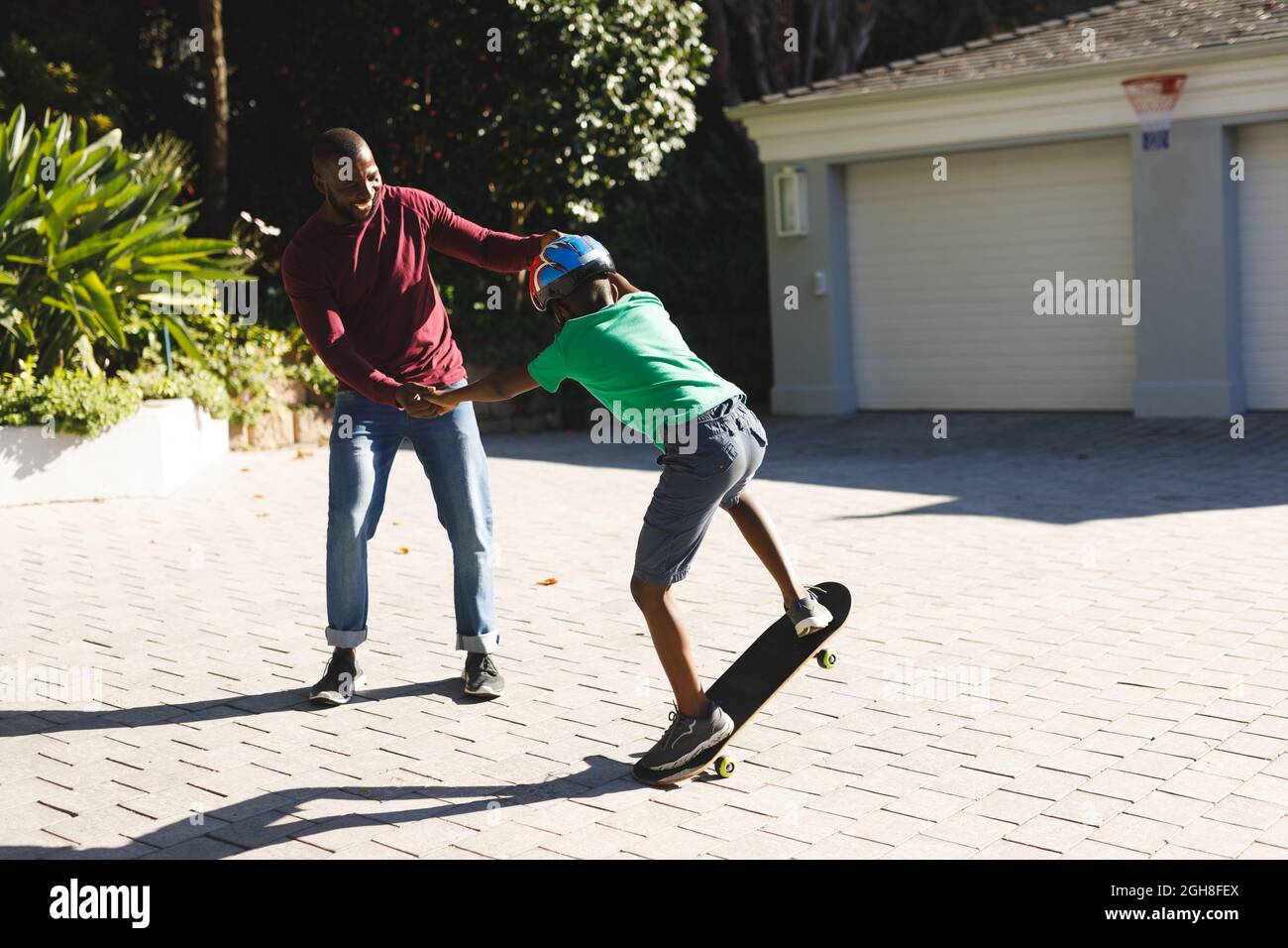 Père afro-américain souriant et aidant son fils à s'équilibrer sur le skateboard dans le jardin Banque D'Images