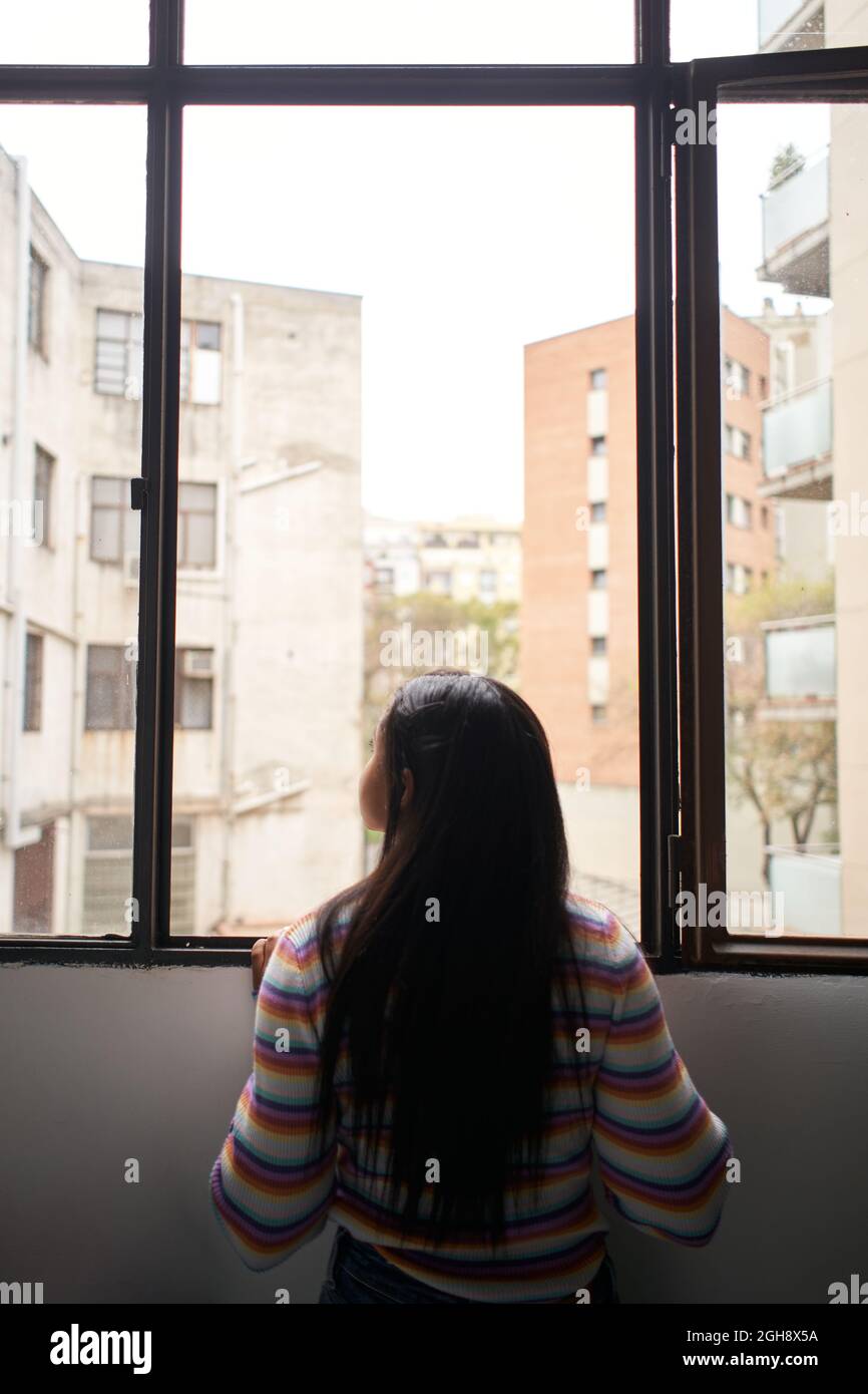 Photo verticale. Vue arrière une fille africaine s'est penchée hors de la fenêtre, regardant à l'extérieur où il y a de grands bâtiments. Banque D'Images