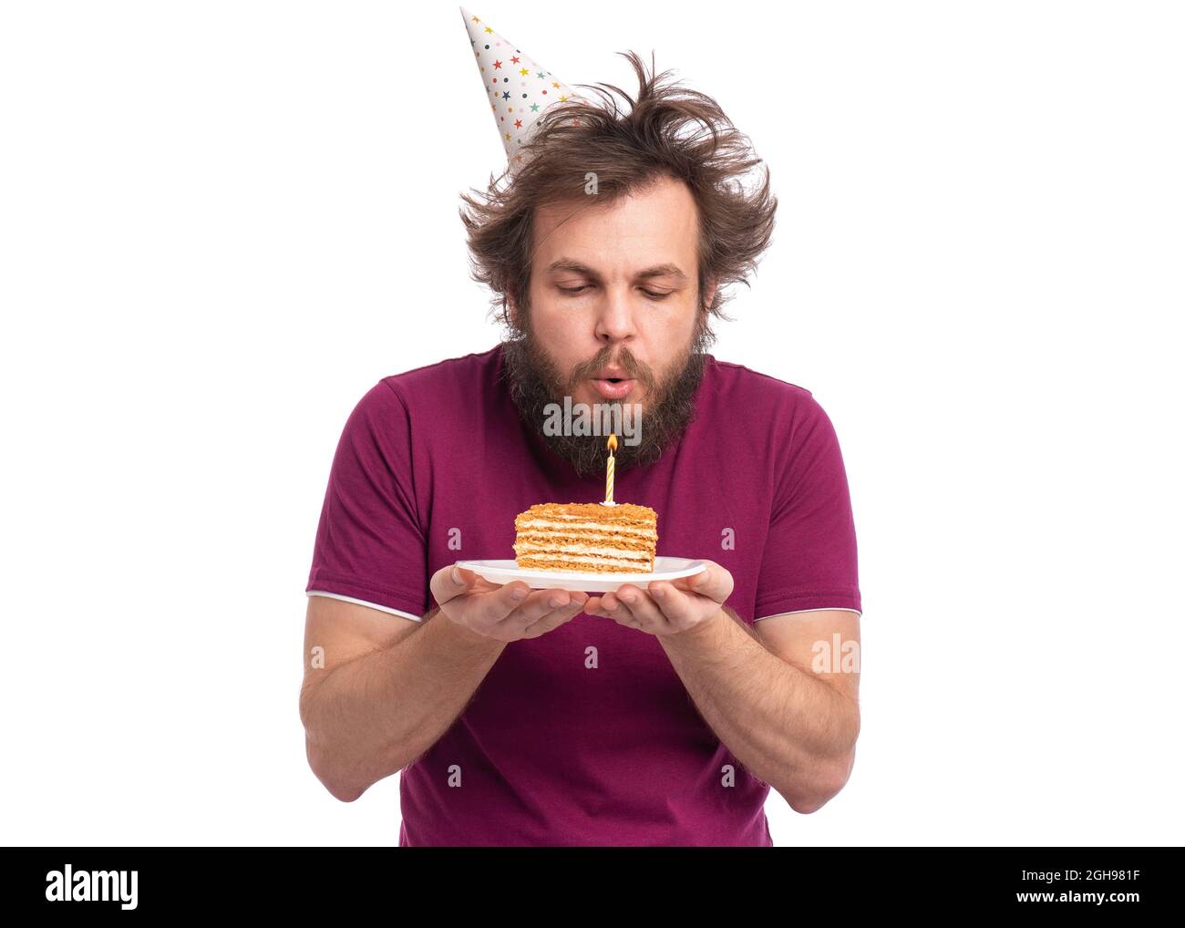 Homme barbu fou avec une coupe de cheveux drôle dans la casquette d'anniversaire, isolé sur fond blanc. Guy soufflant bougie sur son gâteau d'anniversaire. Concept de vacances. Banque D'Images