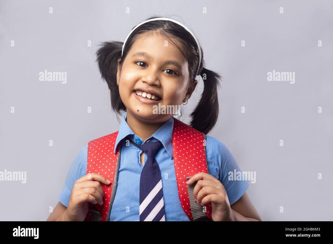 Portrait d'une jeune fille indienne souriante et heureuse en uniforme scolaire bleu avec sac rouge sur fond gris Banque D'Images