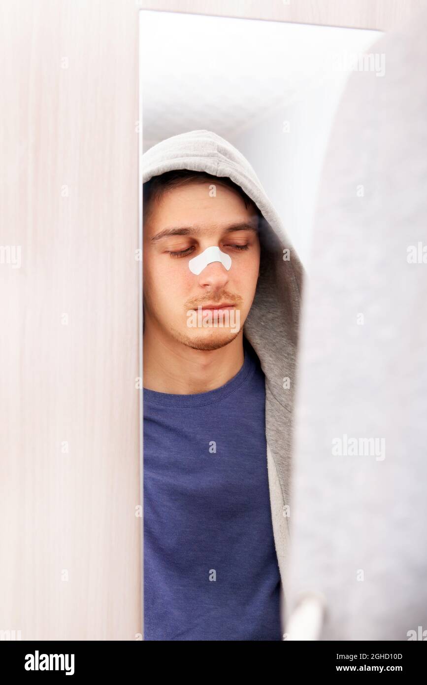 Triste jeune homme avec un plâtre collant sur le nez près du miroir à la maison Banque D'Images