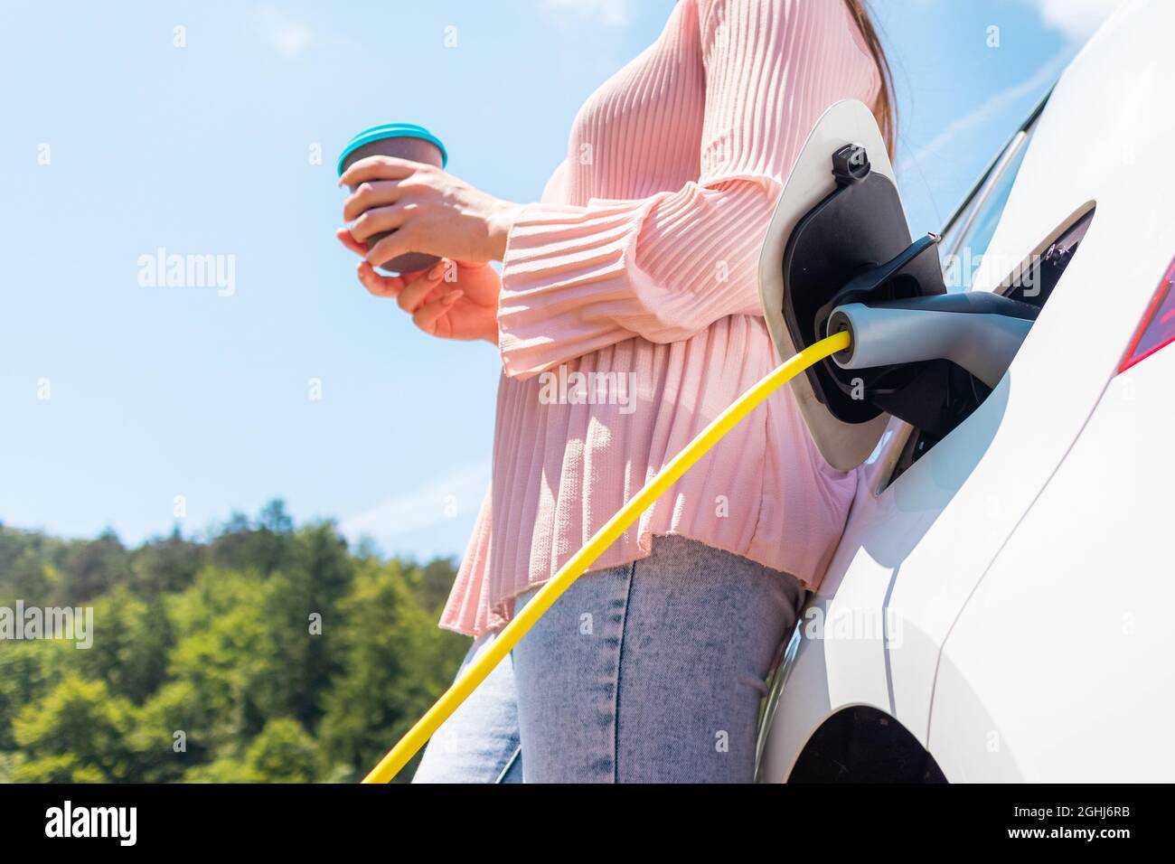 Femme naviguant sur un téléphone mobile et buvant du café tout en attendant de charger sa voiture électrique Banque D'Images
