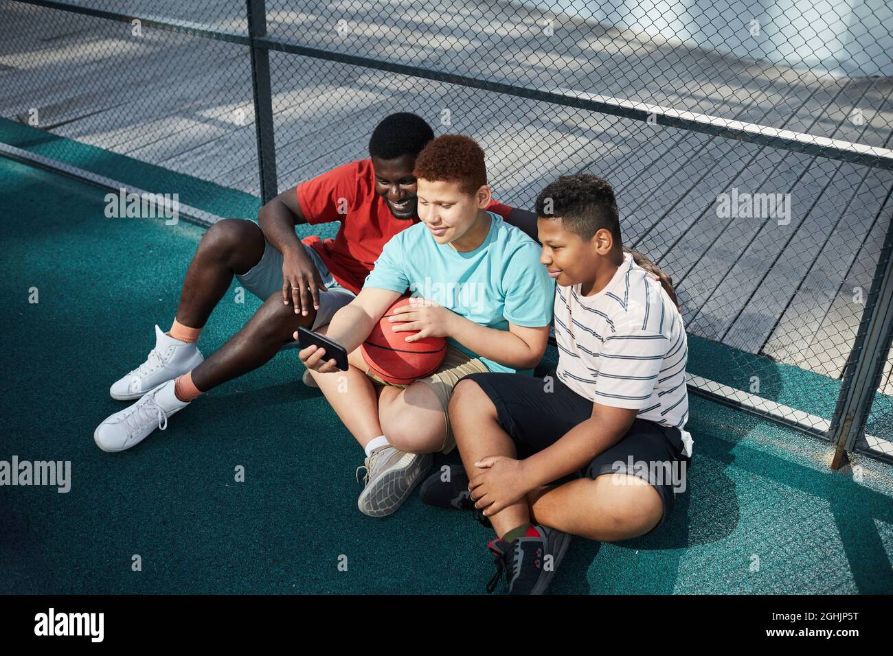 Adolescent assis avec des jambes croisées et un ballon de basket-ball et en utilisant un smartphone tout en posant pour selfie avec des frères sur le terrain de basket-ball Banque D'Images