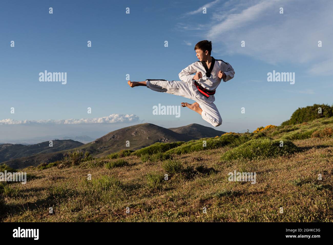 Un garçon de karaté dans un kimono blanc donne des coups de pied dans l'air tout en pratiquant les arts martiaux en plein air. Banque D'Images