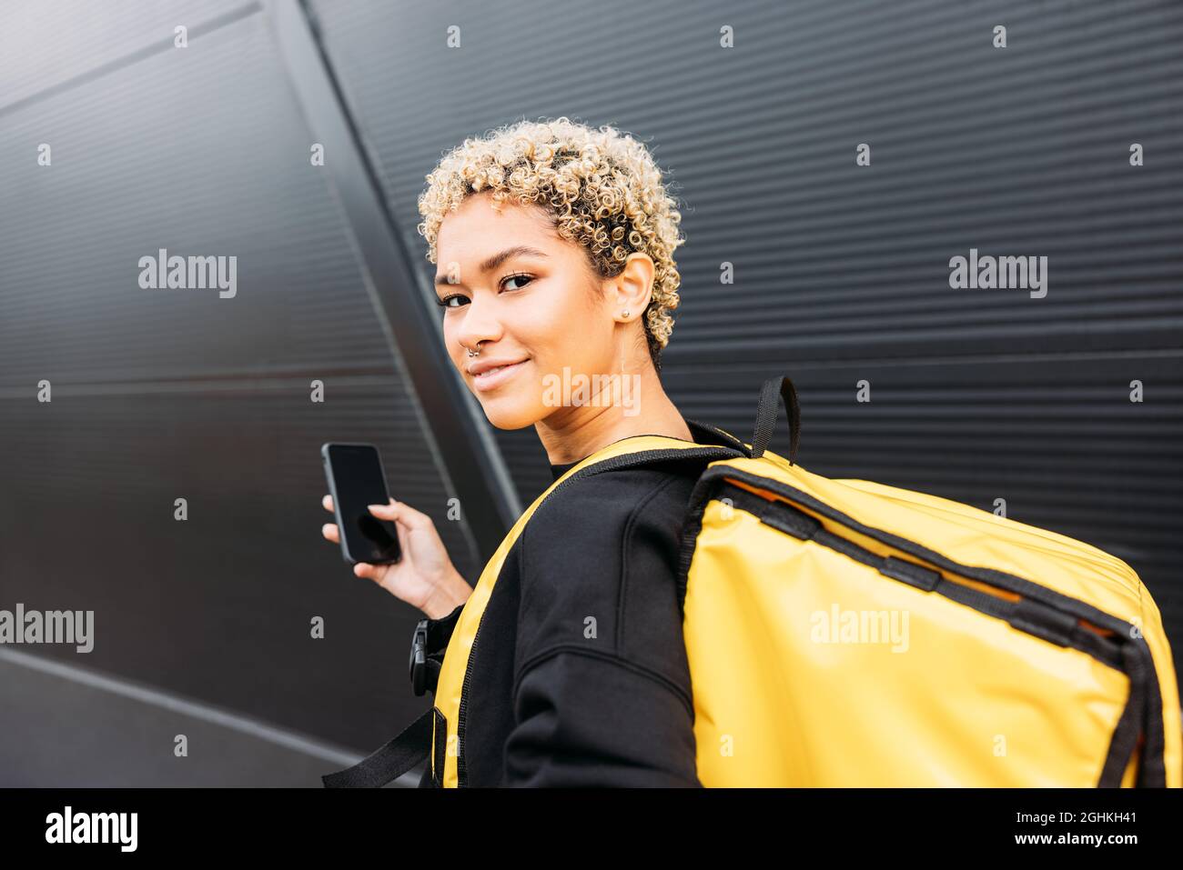 Portrait d'une femme de livraison souriante avec un sac debout sur un trottoir et tenant un smartphone Banque D'Images