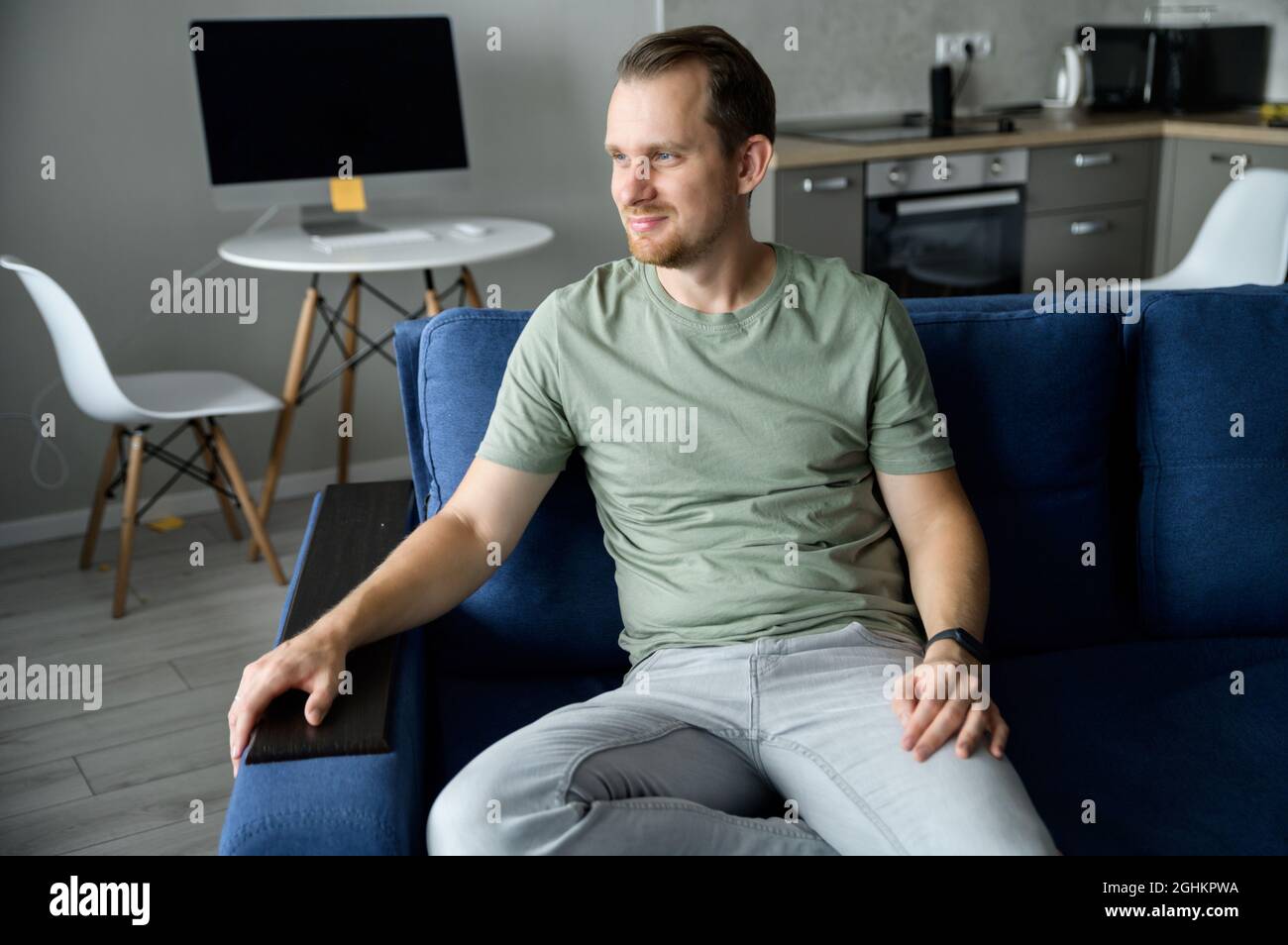 Homme charismatique en tenue décontractée et élégante, assis sur le canapé et regarde loin. Portrait de beau homme serein reposant dans l'appartement moderne. Concept de santé et de bien-être Banque D'Images