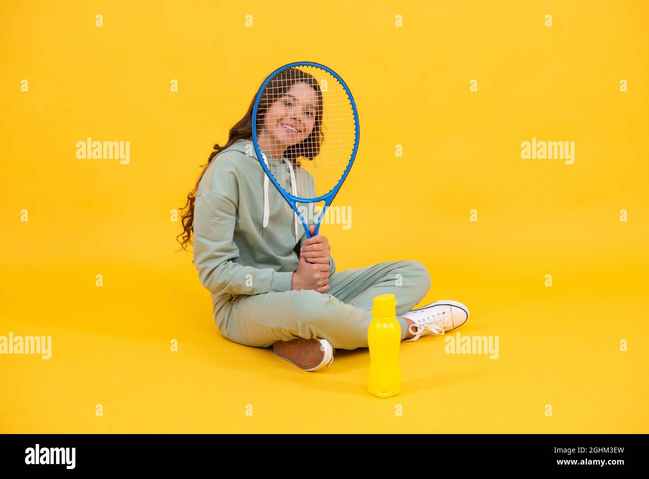 un enfant heureux s'assoit dans des vêtements de sport avec une raquette de squash et une bouteille d'eau sur fond jaune, enfant Banque D'Images