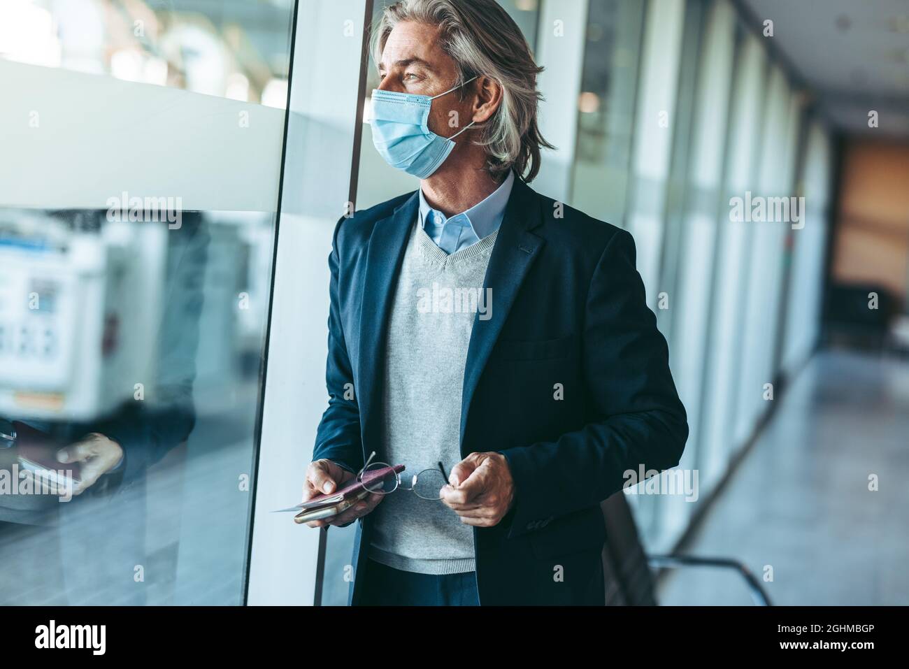 Le voyageur d'affaires porte un masque facial dans le salon d'attente de l'aéroport et regarde à l'extérieur de la fenêtre. Homme d'affaires attendant son vol pendant coron Banque D'Images