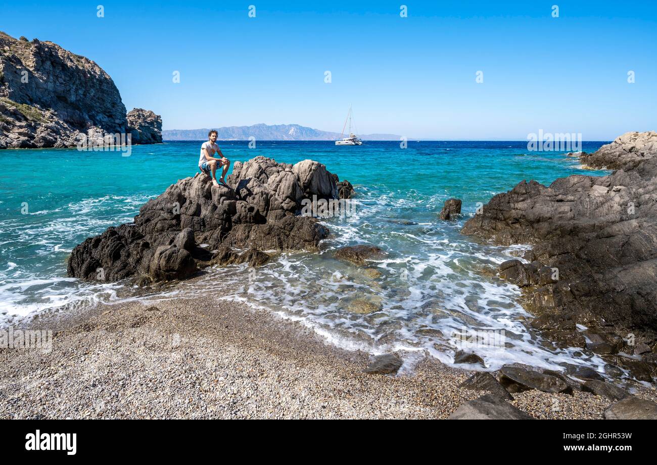 Jeune homme assis sur un rocher, plage avec des roches volcaniques, mer turquoise, catamaran derrière, Gyali, Dodécanèse, Grèce Banque D'Images