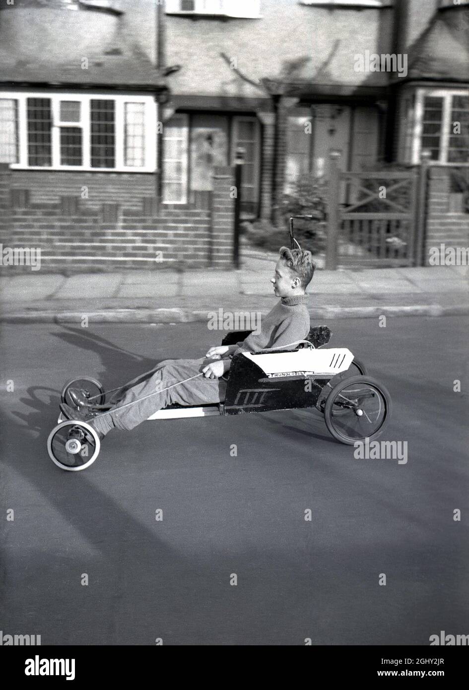 Dans cette photo historique du début des années 1960, un adolescent qui a gagné un chandail à col roulé en laine assis sur un karting fait maison le long d'une route de banlieue, Angleterre, Royaume-Uni. Fabriqué à partir de vieux morceaux de bois fixés à des roues de pram et dirigé avec un peu de corde, beaucoup d'un jeune s'est beaucoup amusé avec une telle machine, qui étaient populaires à la fin des années 50/début des années 60, quand ils étaient communément connus sous le nom de «trolleys». Banque D'Images