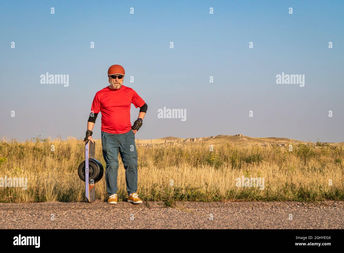 homme senior mature avec un skateboard électrique à une roue sur une route de prairie, paysage de la fin de l'été avec brume et fumée provenant de feux de forêt éloignés Banque D'Images