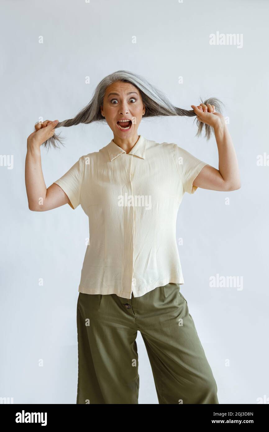 La femme asiatique choquée tient des verrous de cheveux de bonnier sur fond gris clair Banque D'Images