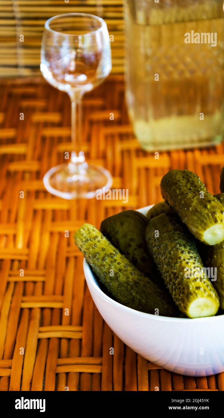 Des concombres marinés dans un petit support de bol blanc sur une table en osier de vigne. En arrière-plan, un petit verre vide pour l'alcool sur un fond de roseaux. Photo verticale. Banque D'Images