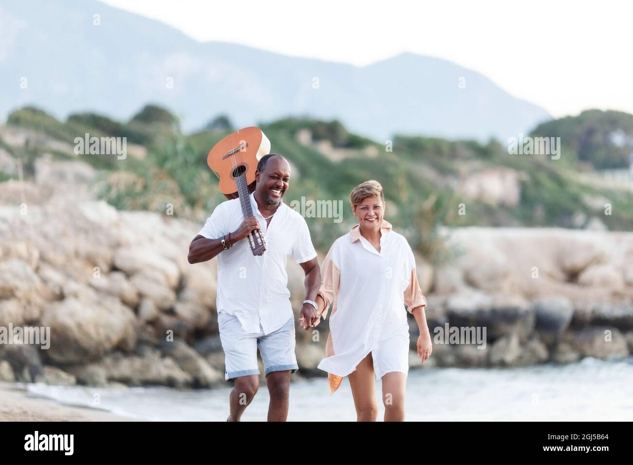 Bonne course mixte, couple d'âge moyen embrassant tout en marchant sur la plage tenant une guitare. Homme tenant guitare avec femme. Banque D'Images Bonne course mixte, couple d'âge moyen embrassant tout en marchant sur la plage tenant une guitare. Homme tenant guitare avec femme. Banque D'Images