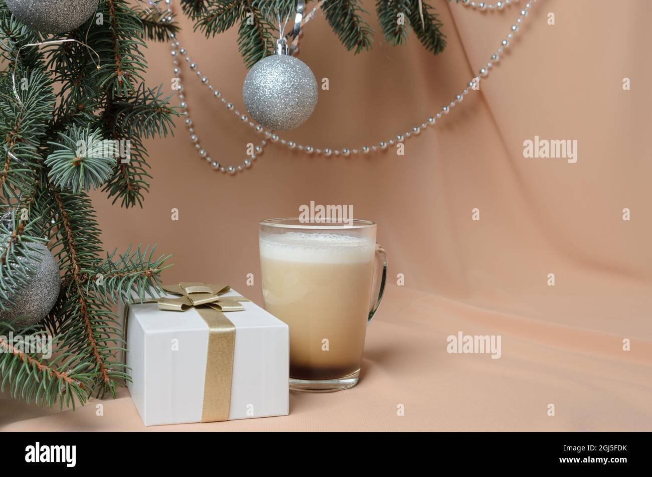 Arbre de Noël, décoré de boules d'argent, d'une boîte cadeau, d'une tasse de verre avec café et lait sur fond de vagues de couleur chair. Mise au point sélective. Banque D'Images