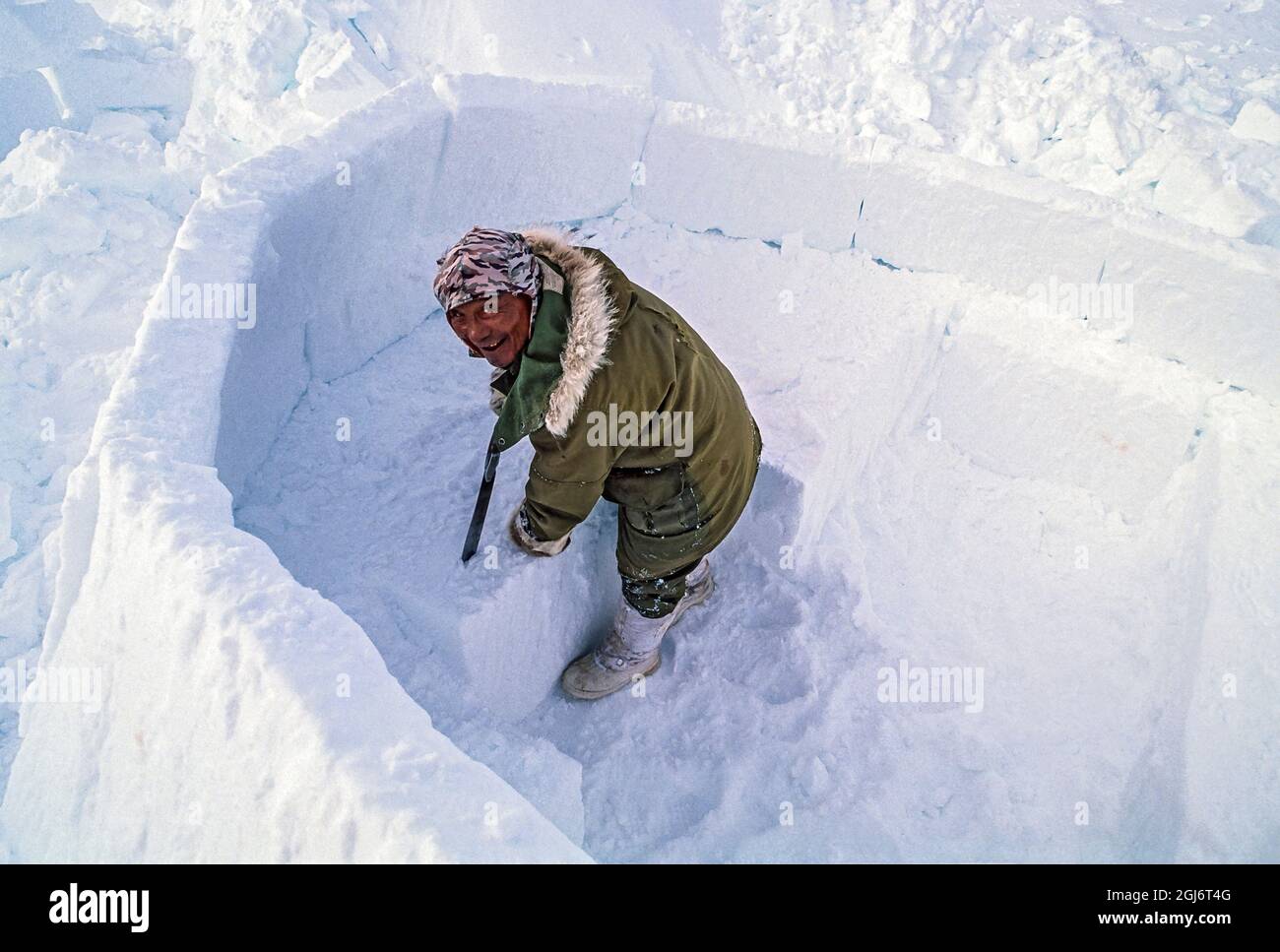 Baker Lake, Nunavut, Canada. Homme aîné inuit, habillé de vêtements modernes d'hiver, coupant des blocs de glace avec un couteau et les tailler tout en construisant le commerce Banque D'Images