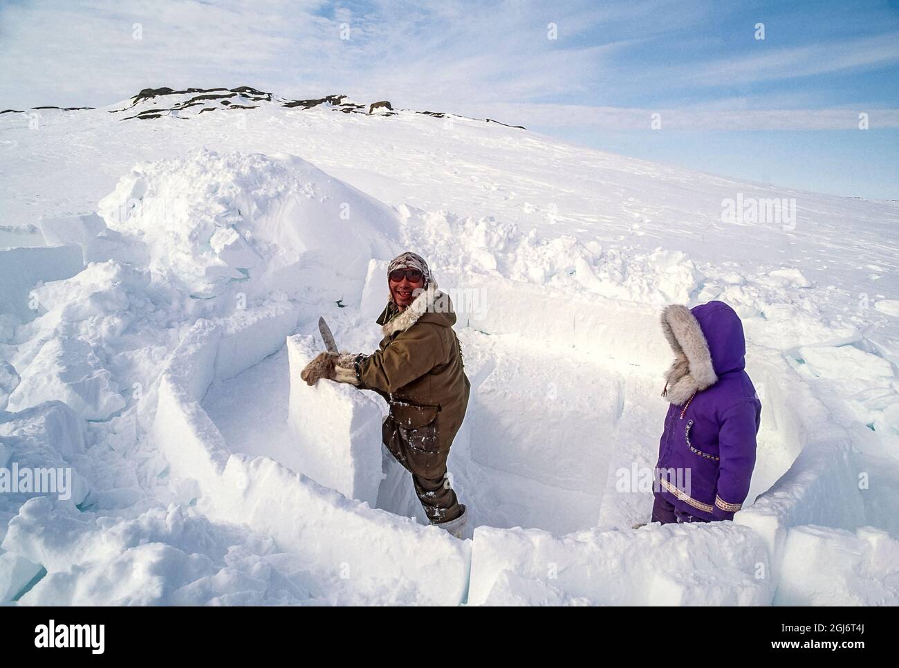 Baker Lake, Nunavut, Canada. Homme aîné inuit, habillé de vêtements modernes d'hiver, coupant des blocs de glace avec un couteau et les tailler tout en construisant le commerce Banque D'Images