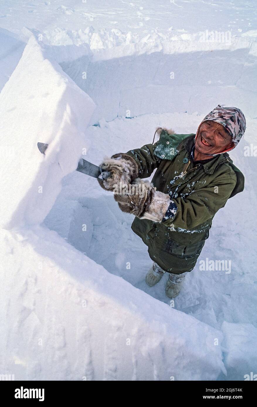 Baker Lake, Nunavut, Canada. Homme aîné inuit, habillé de vêtements modernes d'hiver, coupant des blocs de glace avec un couteau et les tailler tout en construisant le commerce Banque D'Images
