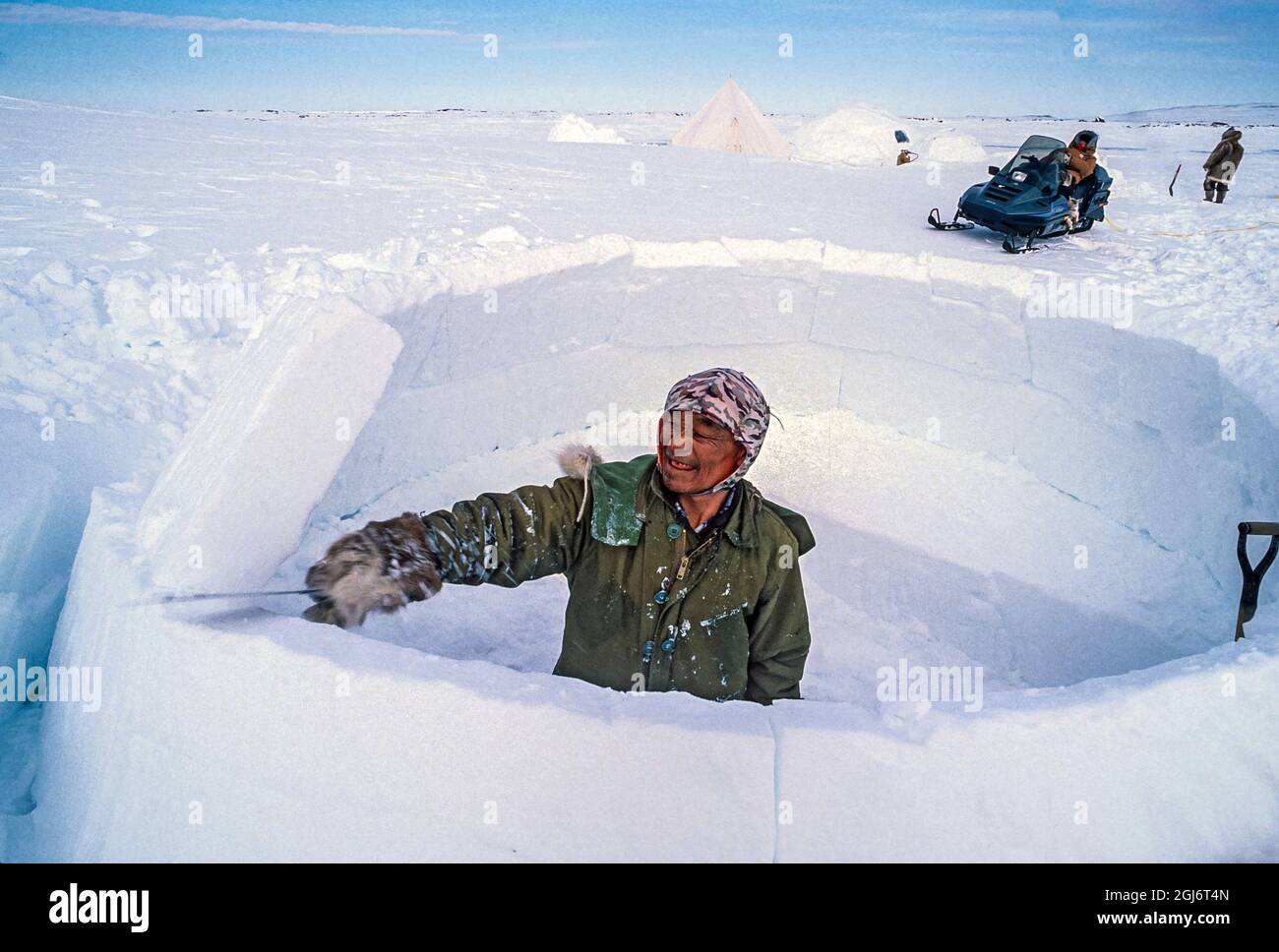 Baker Lake, Nunavut, Canada. Homme aîné inuit, habillé de vêtements modernes d'hiver, coupant des blocs de glace avec un couteau et les tailler tout en construisant le commerce Banque D'Images