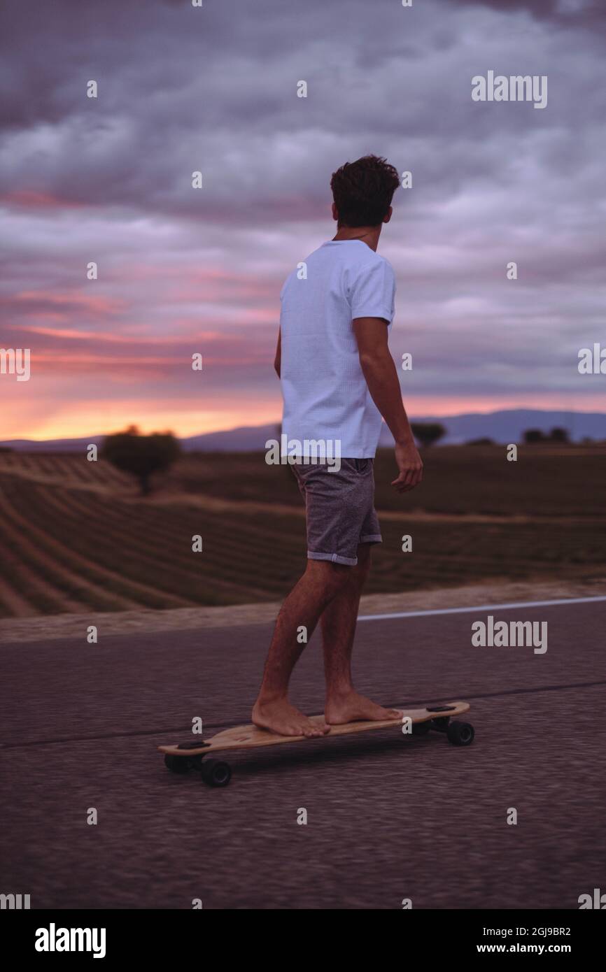 Jeune garçon actif à bord d'un skateboard sur la route et en admirant le  paysage pittoresque au coucher du soleil dans un ciel nuageux. Garçon  sportif, skate et look Photo Stock -