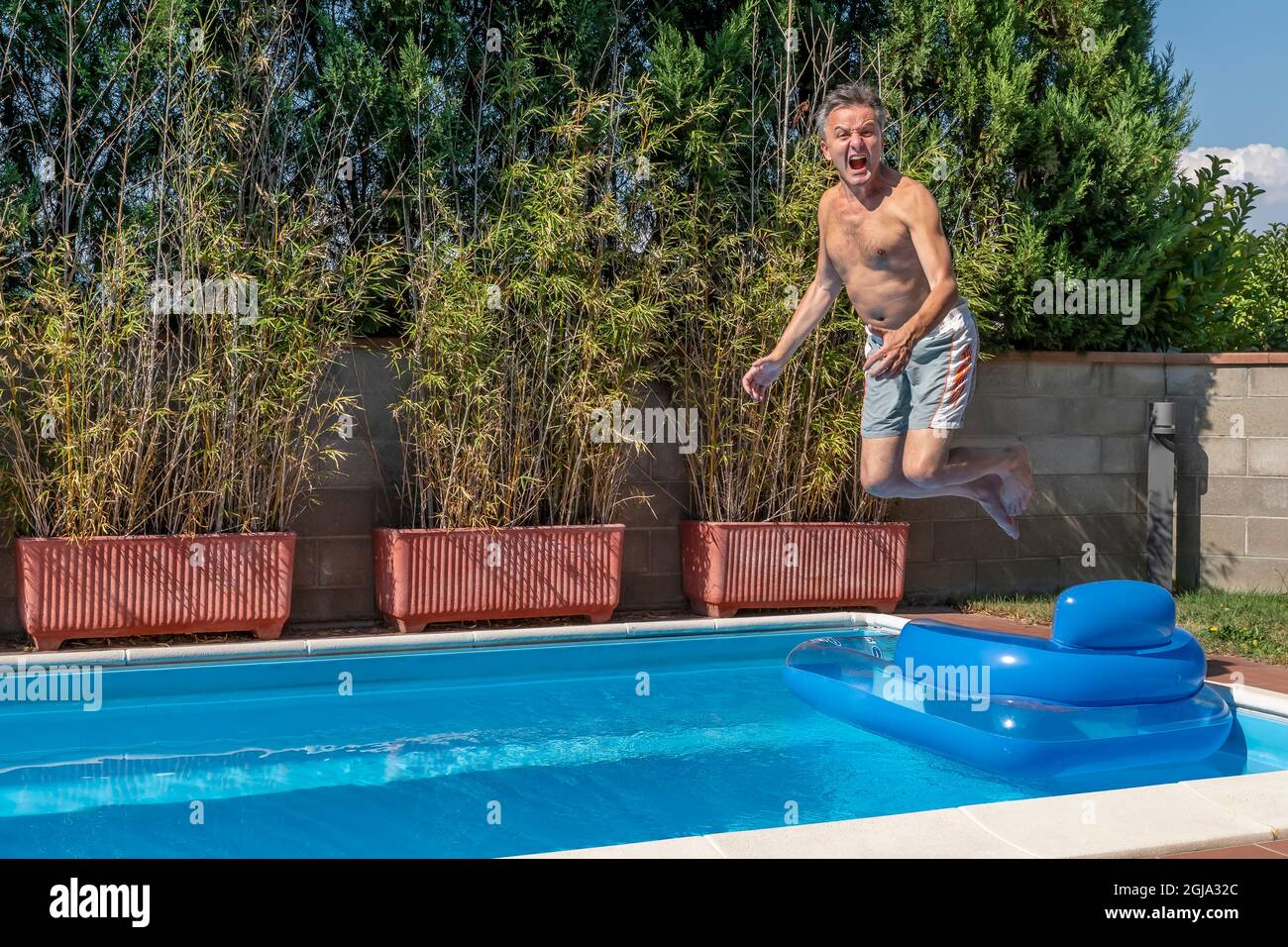 Un homme blanc d'âge moyen plonge dans une piscine, dans un style canonnball, tout en hurlant comme un fou et en sautant au-dessus d'un matelas pneumatique Banque D'Images