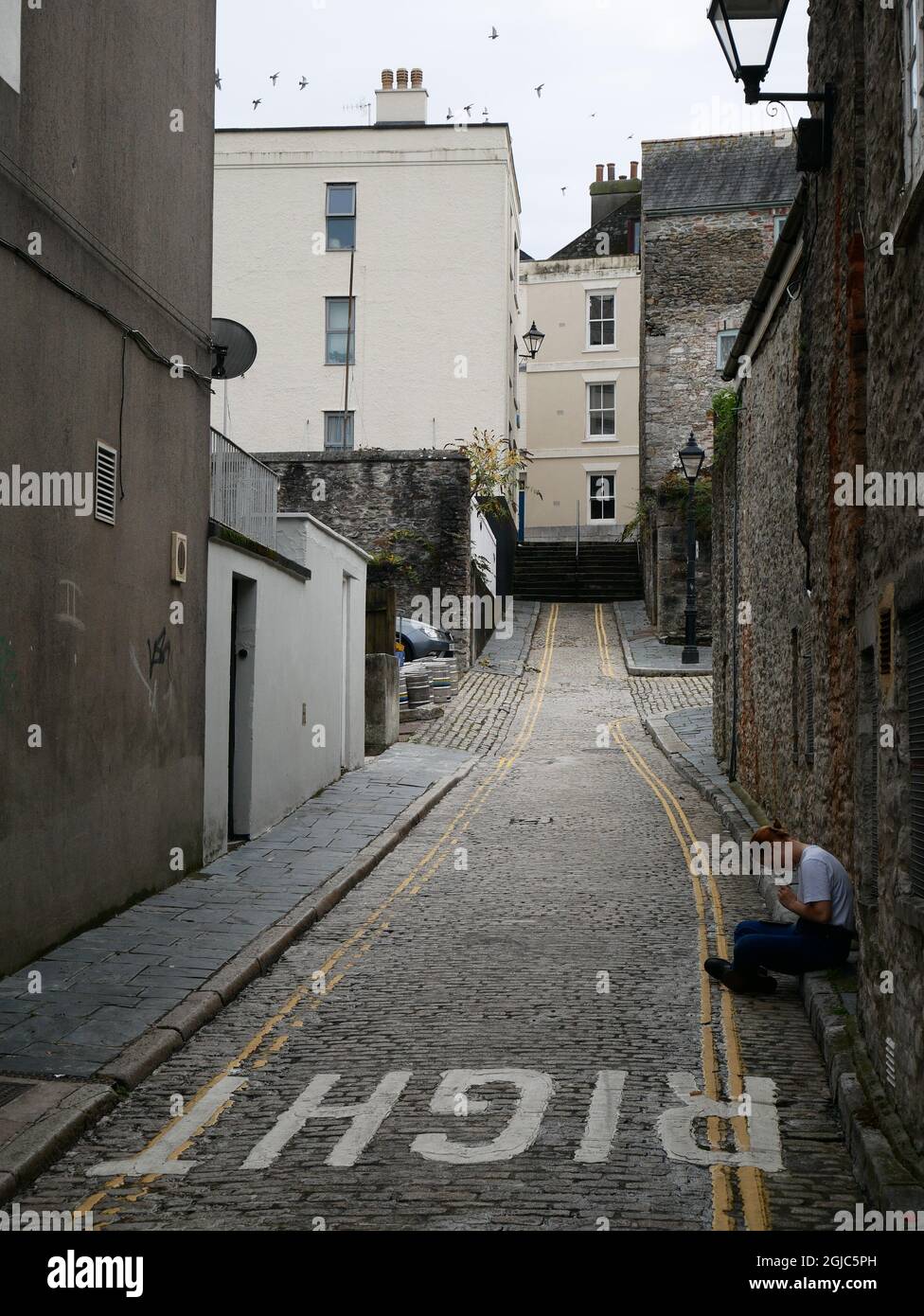 Jeune femme assise seule sur le trottoir dans une petite rue. Barbican, Plymouth, Devon, Royaume-Uni Banque D'Images