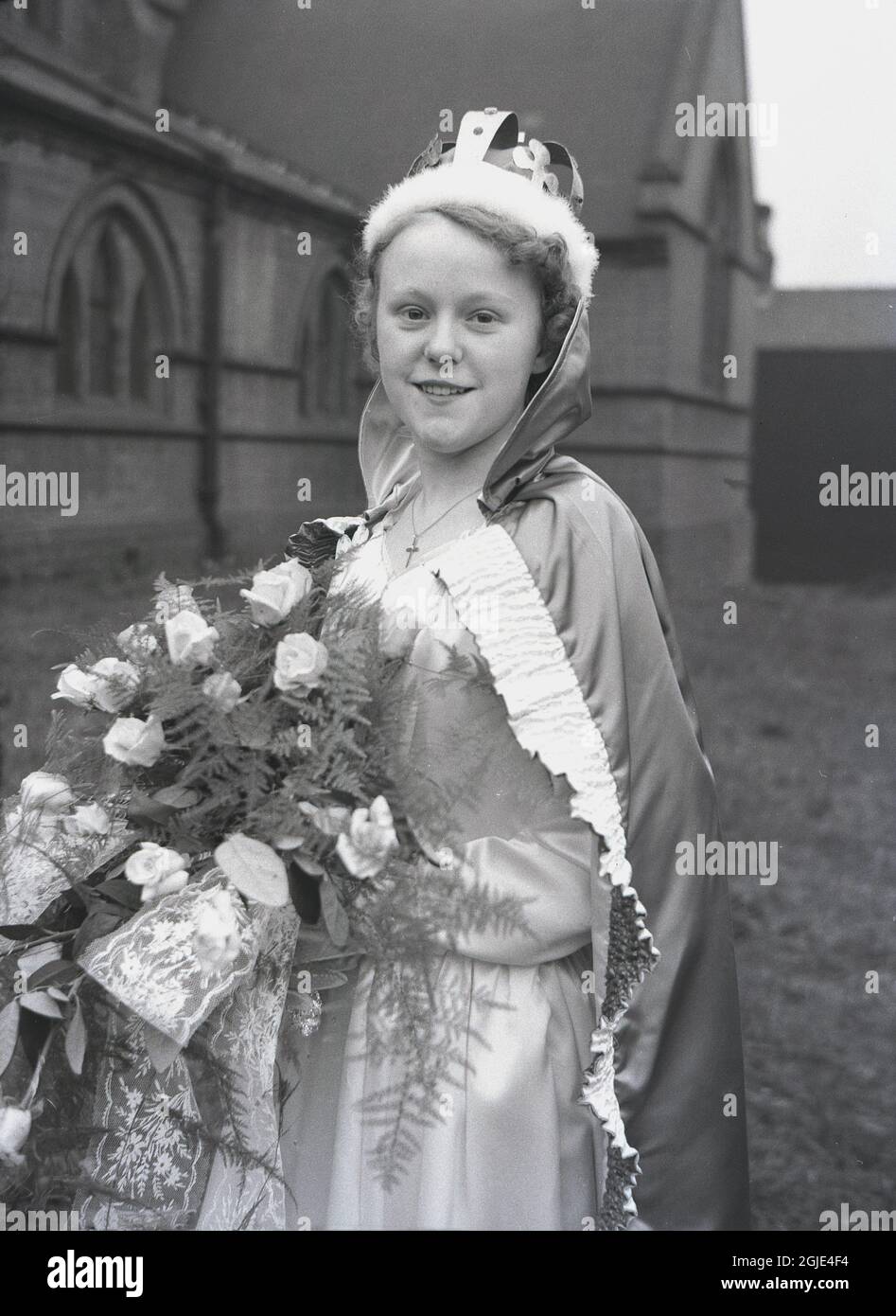 1956, historique, une adolescente, la Reine du jour de mai debout dans le domaine d'une église portant sa couronne, robe, robe et tenant un bouquet de fleurs avant de prendre part au traditionnel carnaval du jour de mai, Leeds, Angleterre, Royaume-Uni. Banque D'Images