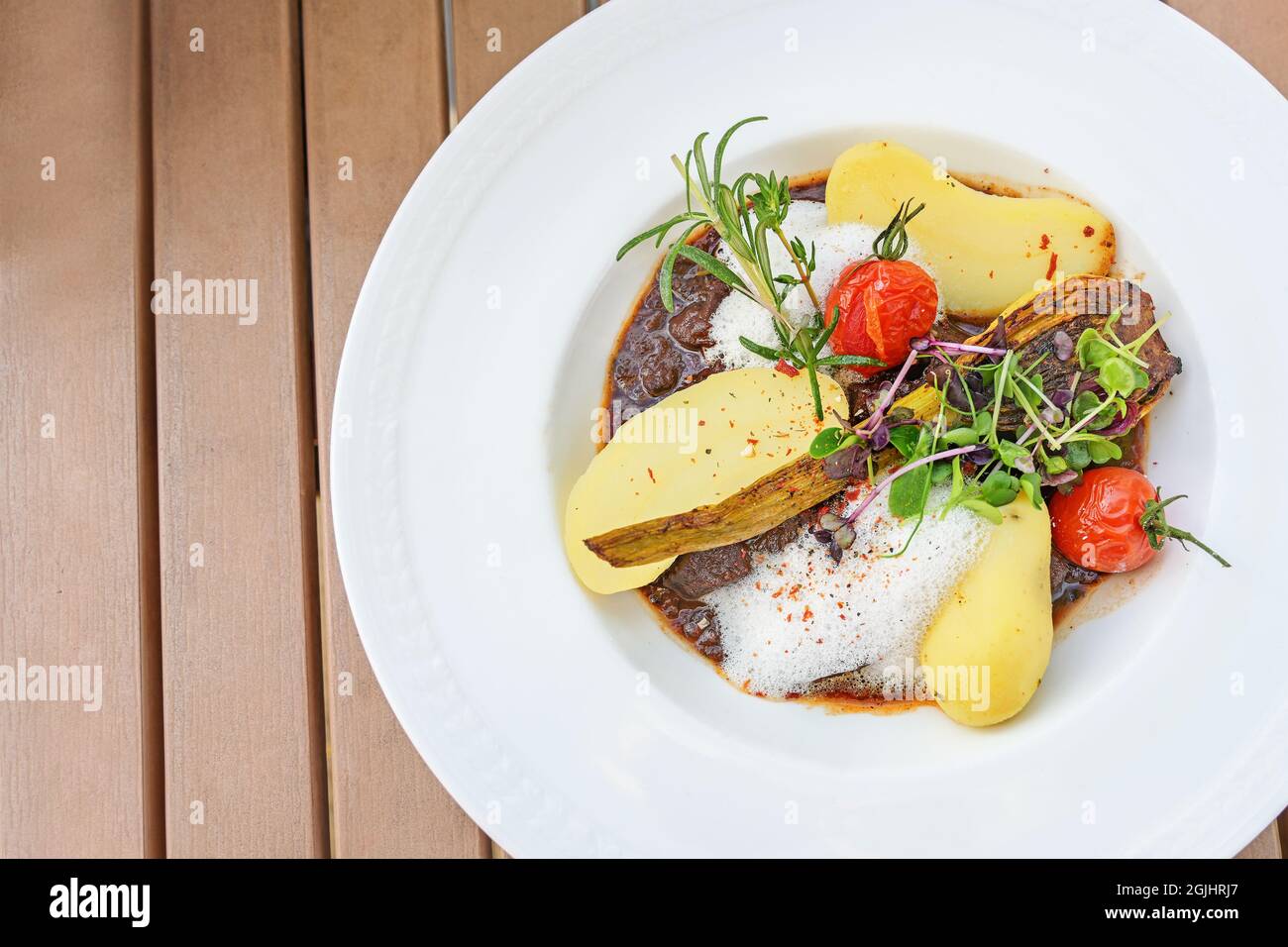 Ragoût de bœuf avec pommes de terre, tomates, fenouil rôti et garniture de cresson, herbes et pousses, servi avec de la mousse décorative sur une assiette blanche sur une ta en bois Banque D'Images