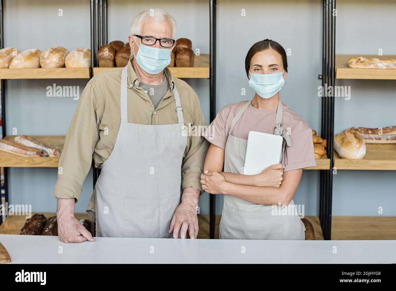 Portrait de boulangers réussis en tabliers et masques debout près du lieu de travail contre présentation avec assortiment de pains Banque D'Images