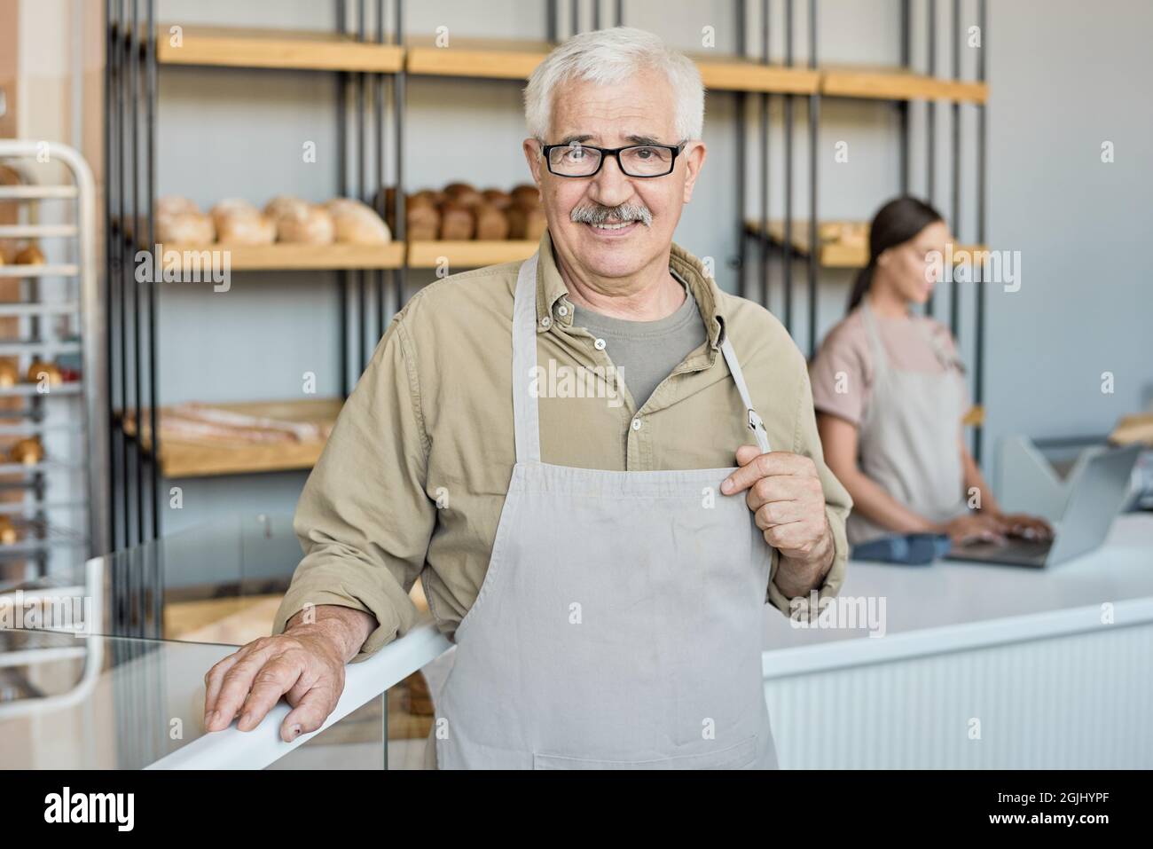 Portrait d'une boulangère vieillie avec moustache tenant un piège de tablier tout en posant contre l'étalage avec assortiment de pain frais Banque D'Images