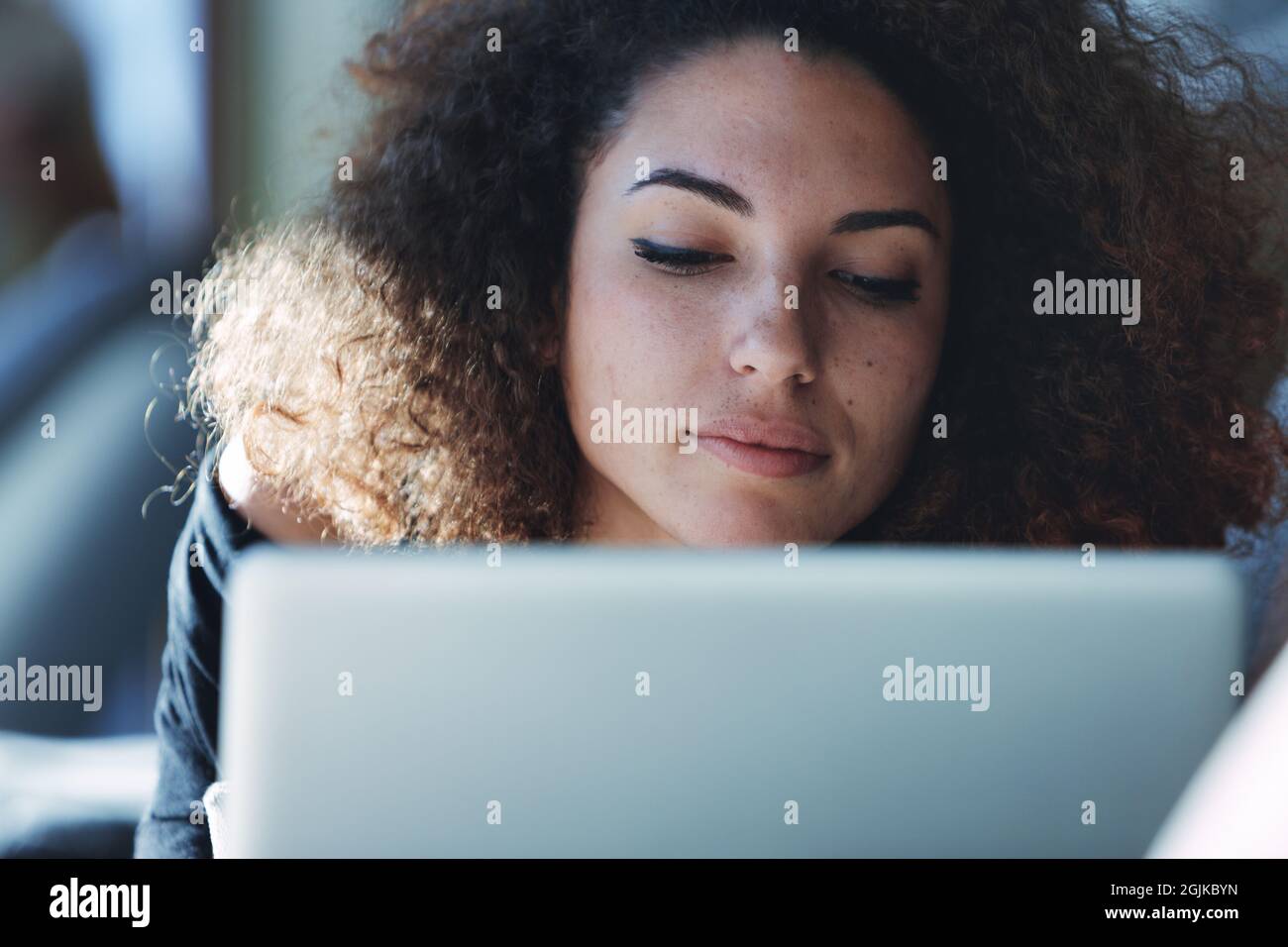 Jeune femme attrayante avec des cheveux bouclés travaillant sur son ordinateur portable en gros plan sur son visage avec une expression engrosée et des yeux détournés Banque D'Images