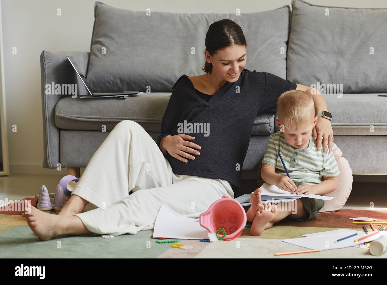 Jeune femme enceinte souriante assise sur le sol et commentant le dessin de son fils dans la salle de séjour Banque D'Images