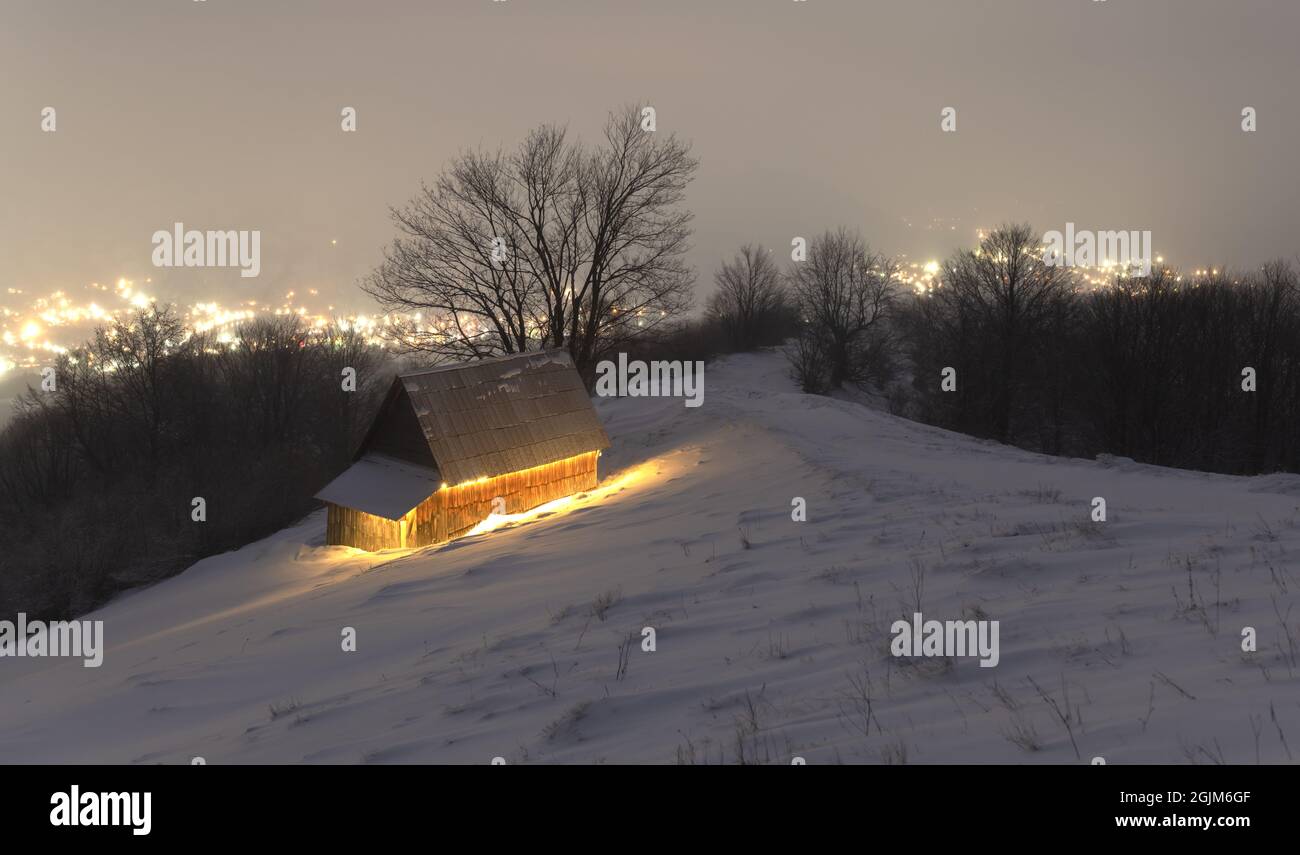 Paysage d'hiver fantastique avec maison en bois lumineux sur fond de lumières de ville dans le brouillard. Chalet confortable dans les montagnes Carpathian. Concept de vacances de Noël Banque D'Images