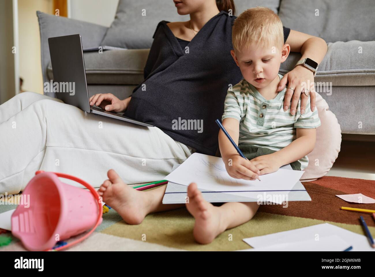 Petit fils indépendant concentré assis sur le sol et de dessiner pendant que sa mère travaille avec un ordinateur portable Banque D'Images