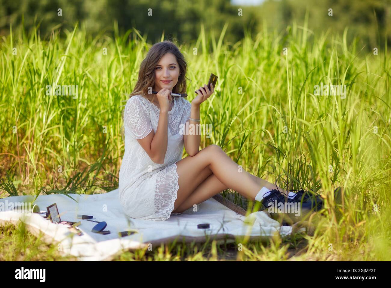 Jeune femme blonde en robe blanche est assise sur une plaque de pique-nique dans une grande herbe. Une maquilleuse féminine avec accessoires de maquillage se répand autour d'elle. Banque D'Images
