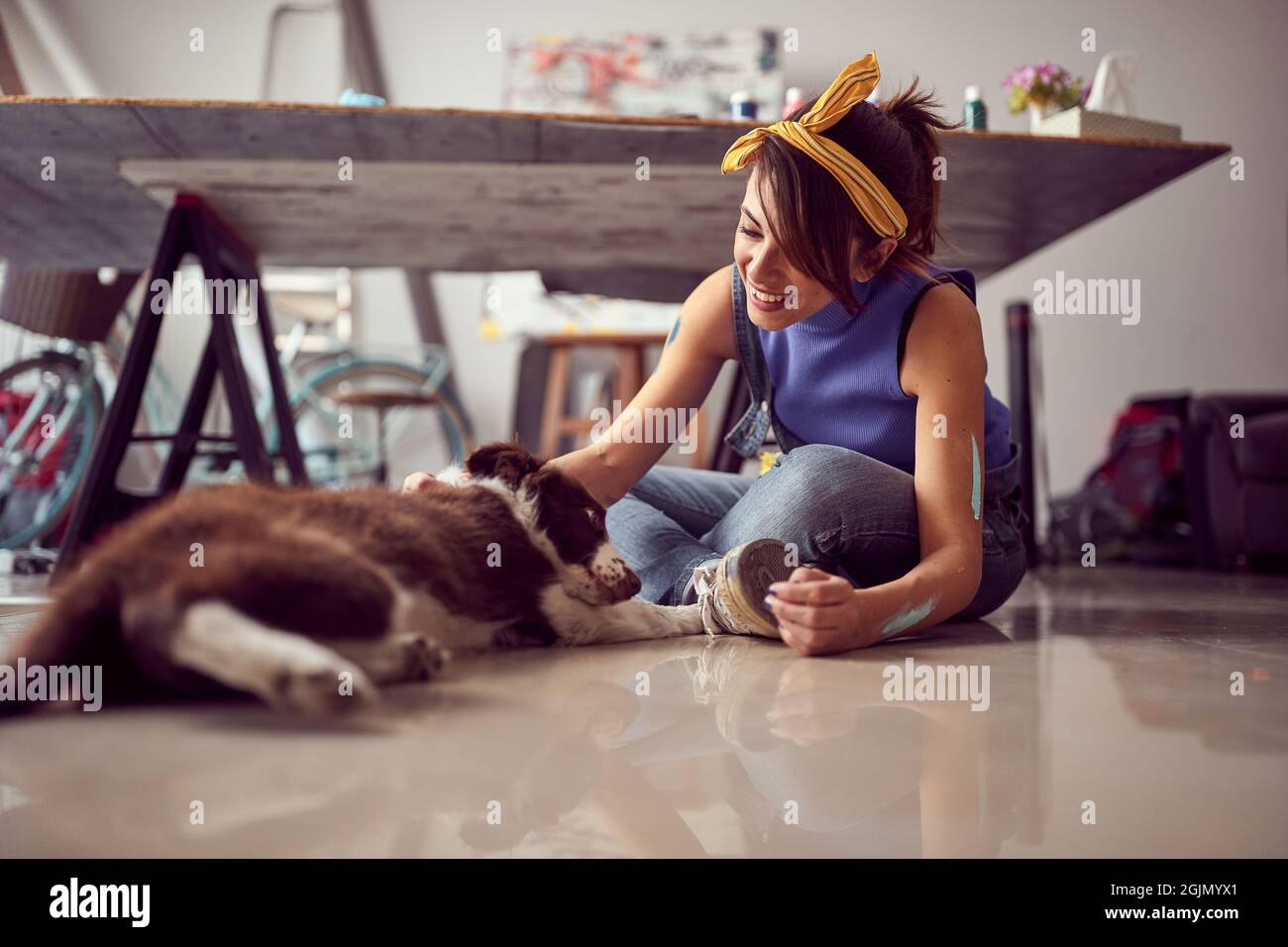 Une jeune artiste est assise sur le sol dans une atmosphère détendue dans le studio et joue avec son chien tout en faisant une pause de travail sur un nouveau p Banque D'Images