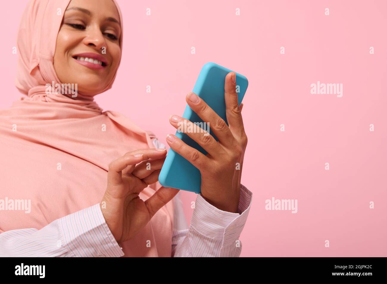 Souriant avec un sourire tortu femme musulmane mature d'origine ethnique du Moyen-Orient dans un hijab rose avec un smartphone dans une couverture bleue sur ses mains, isolée sur p rose Banque D'Images