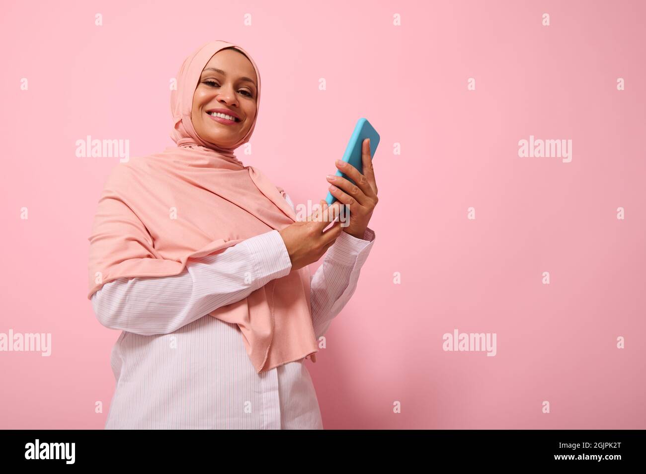 Souriant avec un sourire éclatant une femme musulmane mûre d'origine ethnique du Moyen-Orient dans un hijab coloré avec un smartphone dans la couverture bleue sur les mains, regarde l'appareil photo, est Banque D'Images