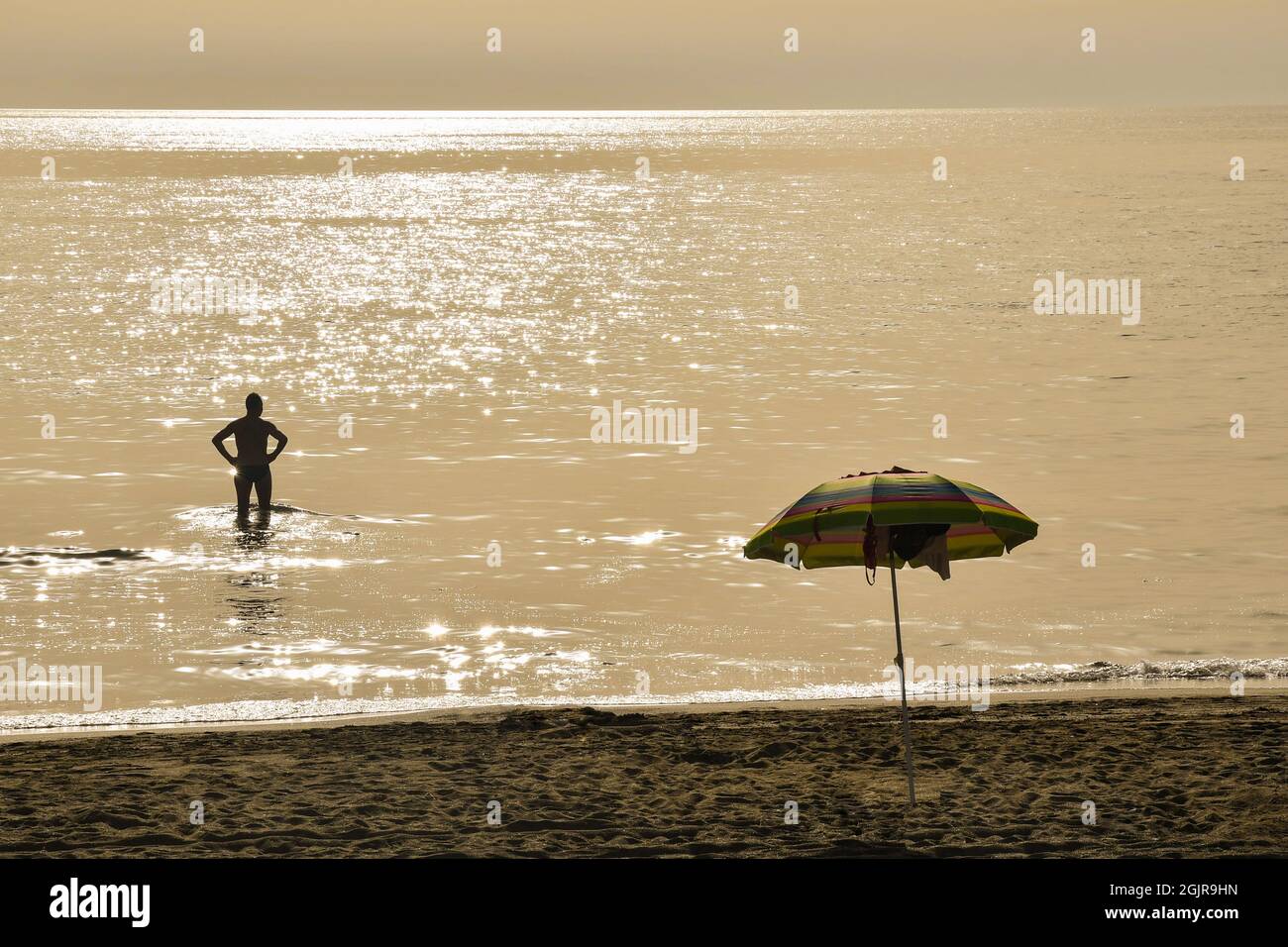 Vue arrière de la silhouette d'un homme debout dans la mer au coucher du soleil avec un parasol sur la plage de sable, Marina di Castagneto Carducci, Toscane Banque D'Images